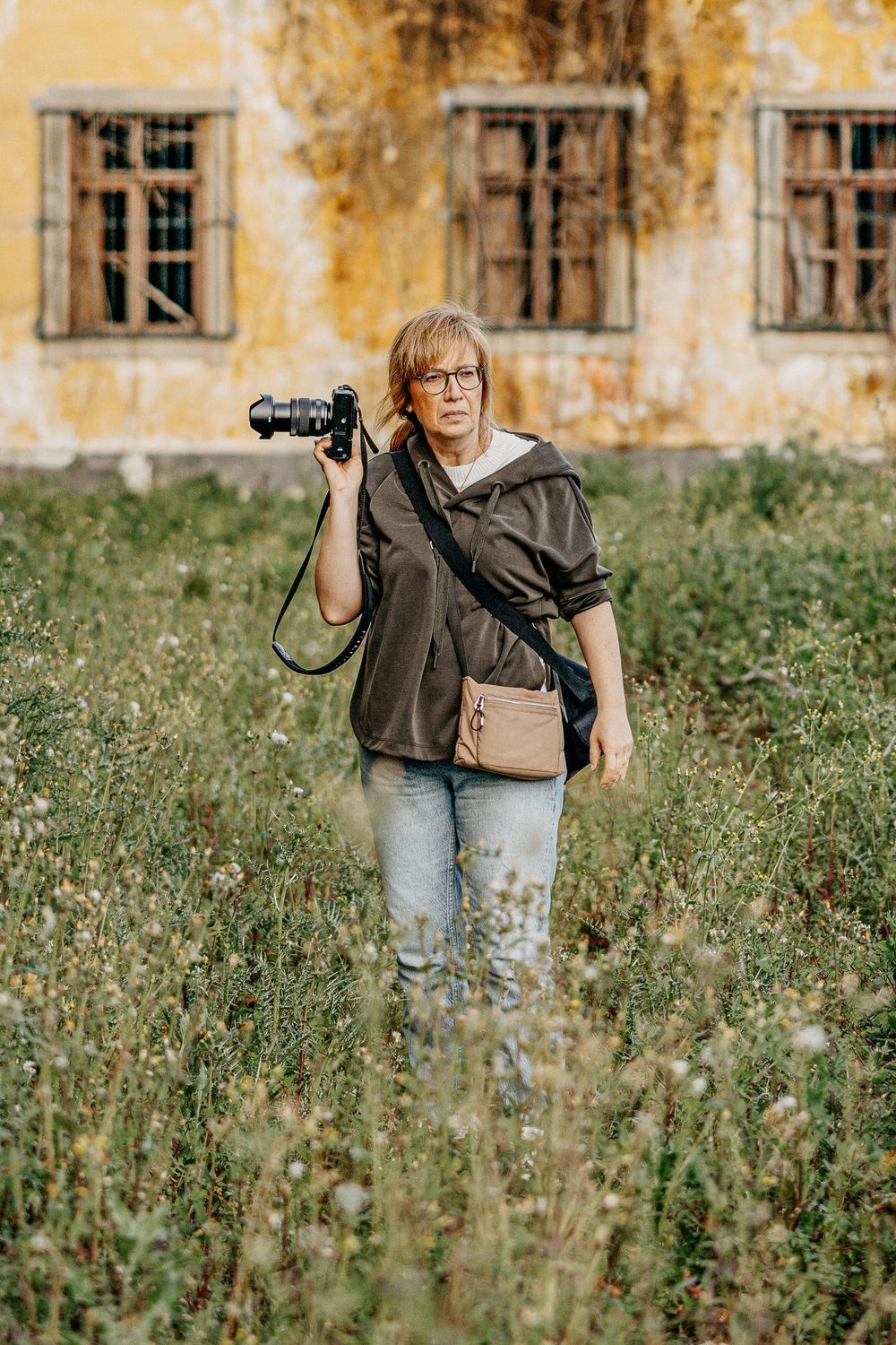 Photographer with camera standing in overgrown field near abandoned building.