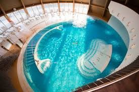 Curved indoor swimming pool with bright turquoise water surrounded by white lounge chairs in a spa setting.