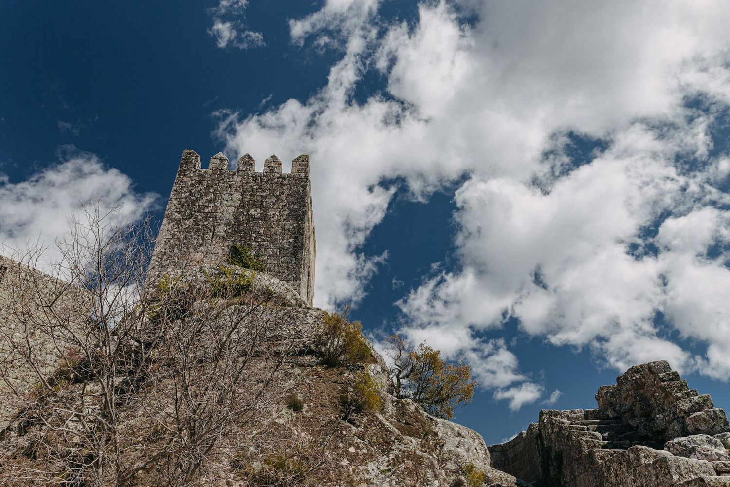 Ancient stone tower rises against a dramatic blue sky with white clouds in a medieval landscape.