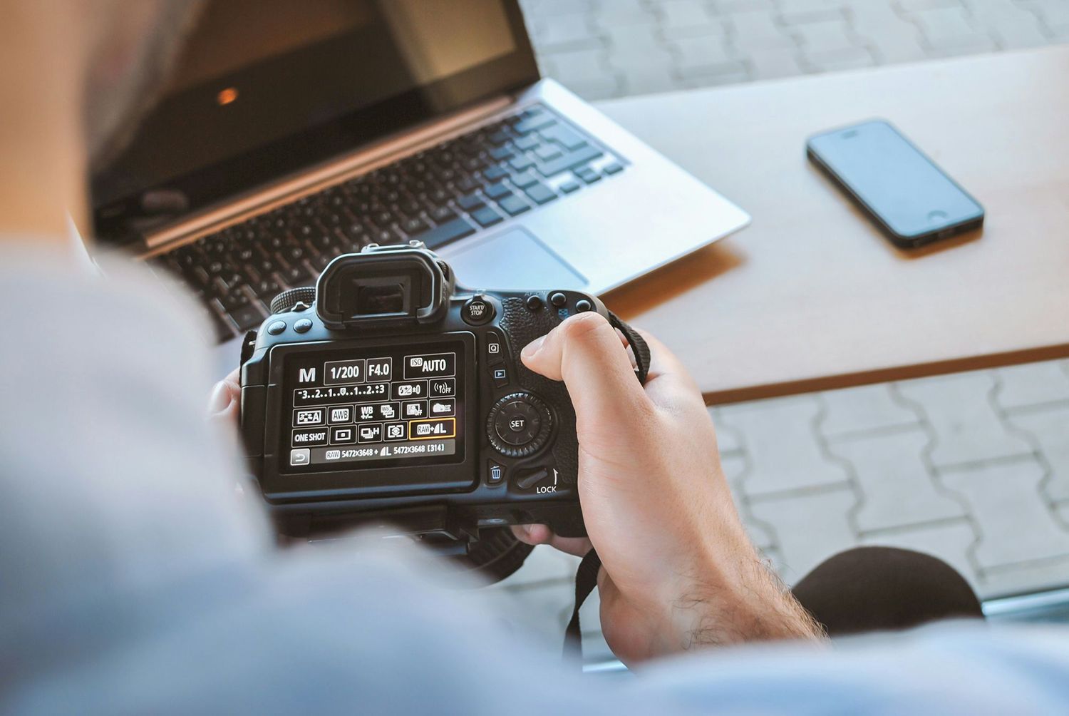 Hands adjusting settings on a DSLR camera with laptop and smartphone on desk.