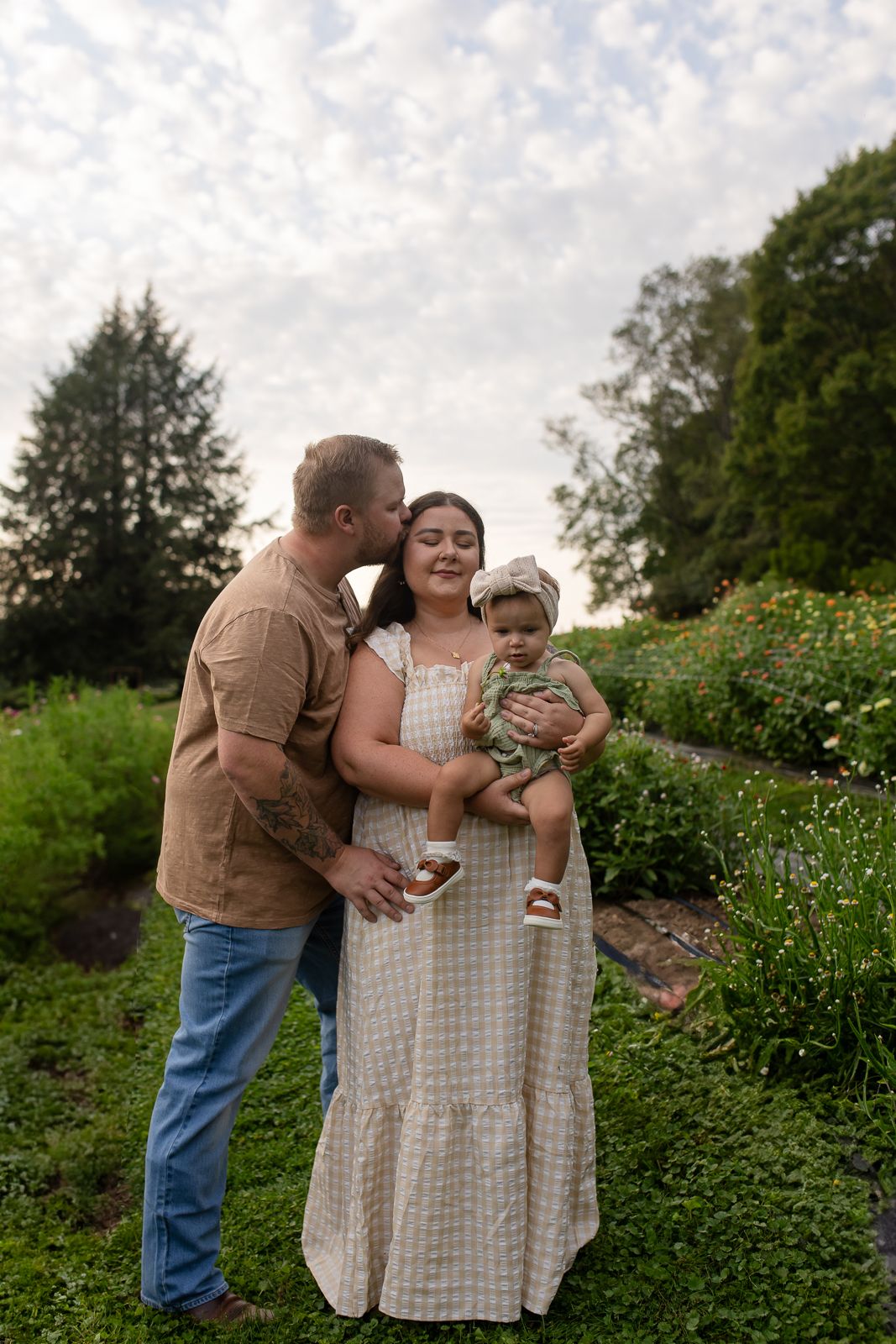 man kissing woman in a gingham dress on the temple while she holds their baby