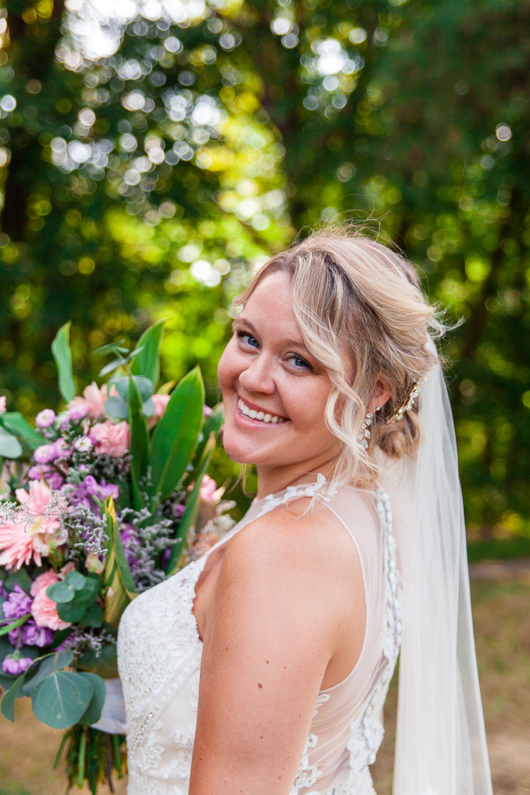 bride smiling happily at the camera while holding bouquet filled with pink and purple flowers and greenery