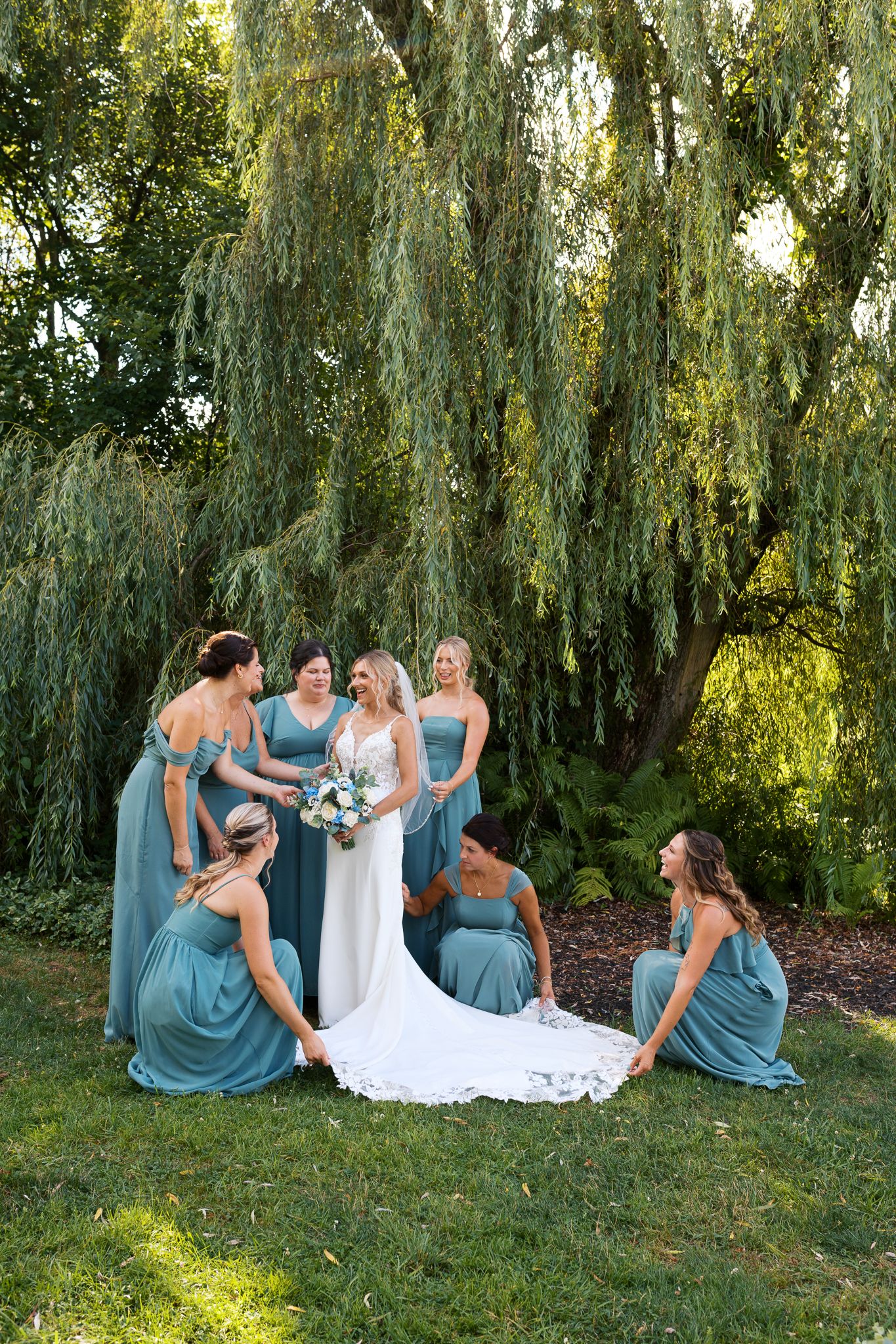 bridesmaids in teal dresses getting the bride ready in her lace wedding gown in front of a weeping willow tree