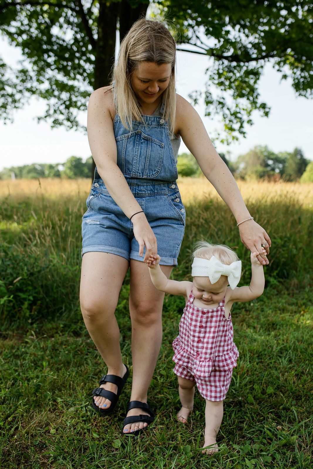 little girl in red gingham outfit and white box holding hands with her mom in denim shorts romper