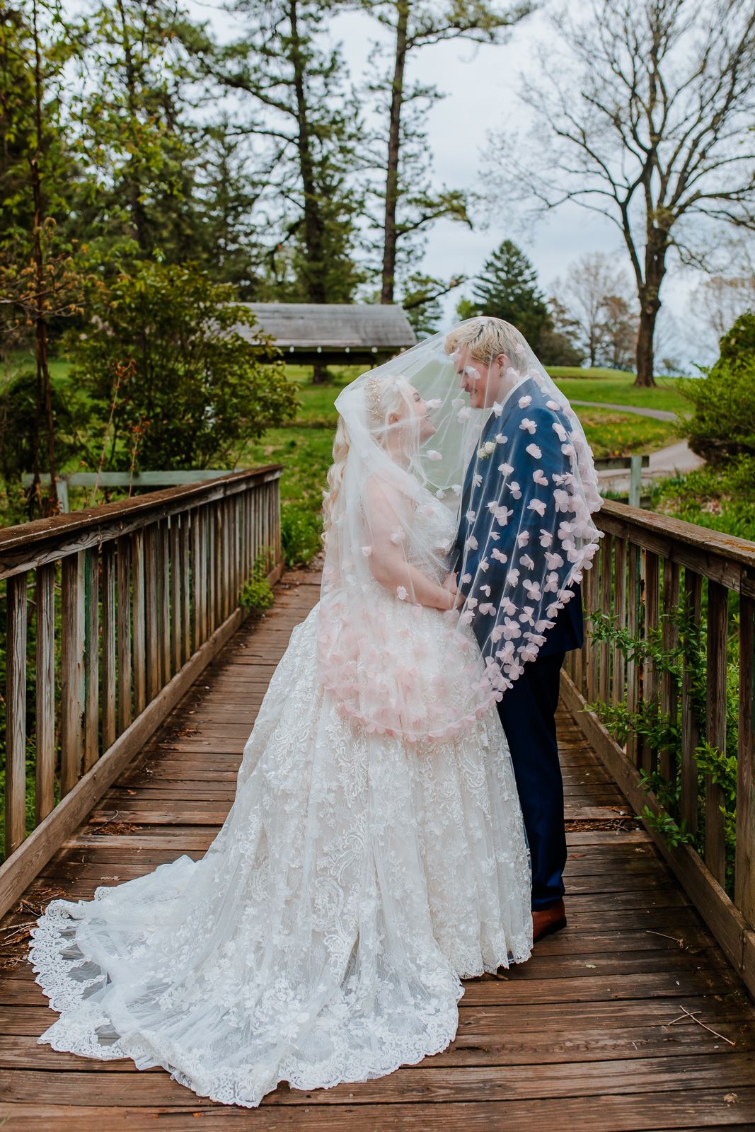 man and woman under a long veil with floral detailing standing on a wooden bridge
