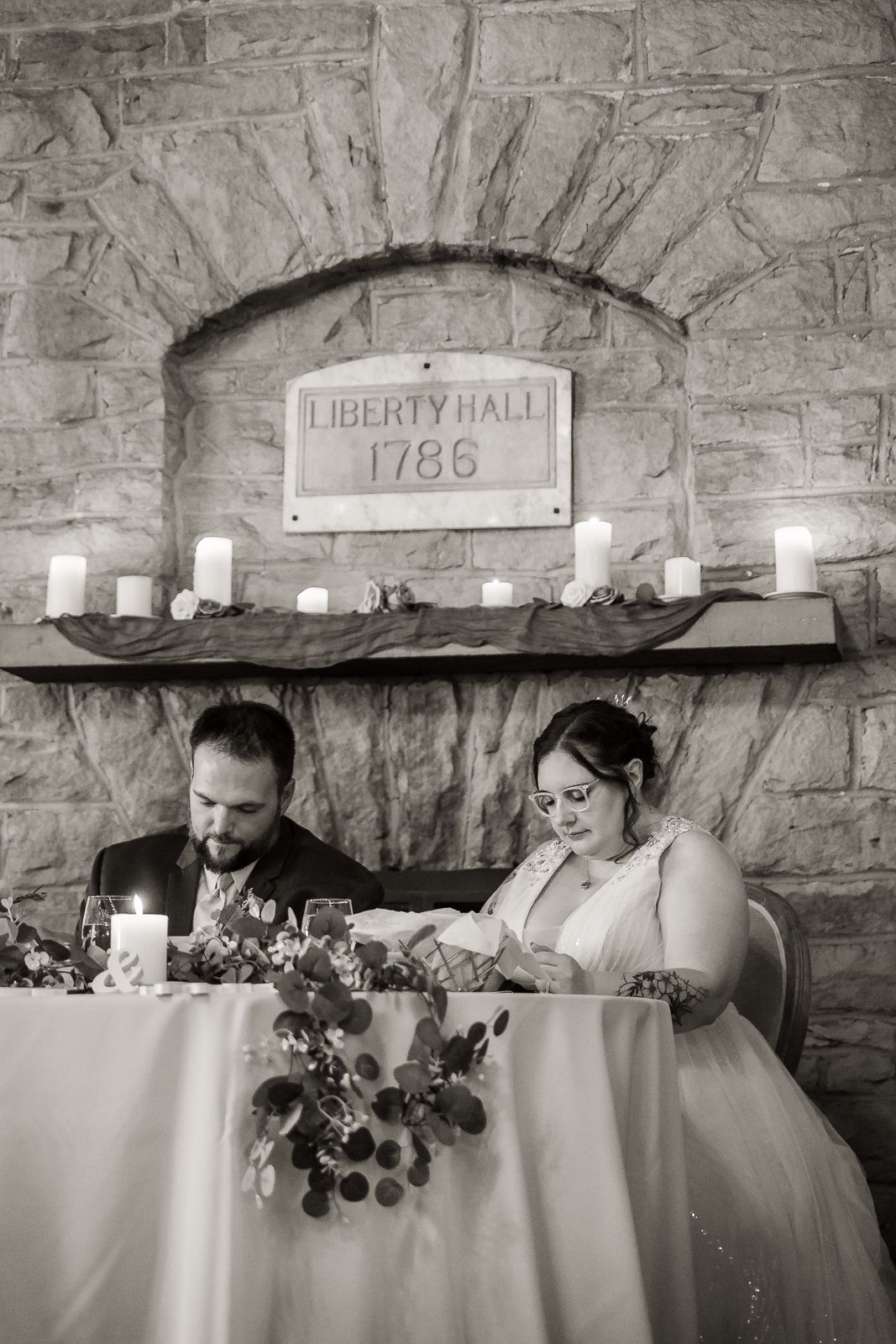 black and white image of bride and groom sitting at sweetheart table in front of stone fireplace reading letters