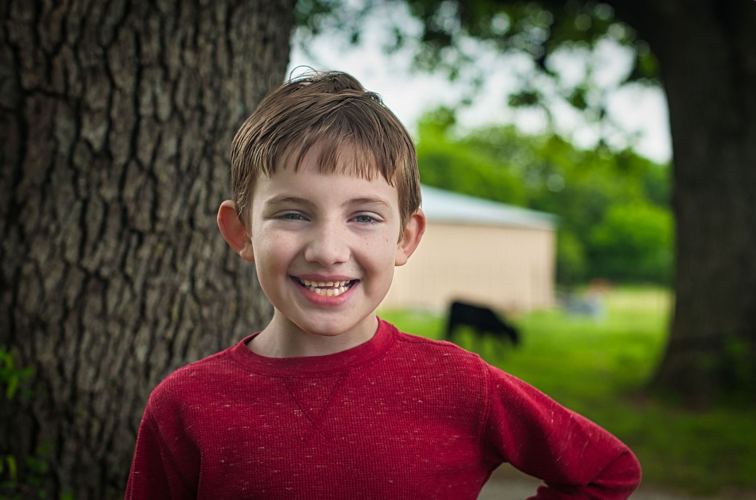 A young person in a red sweater stands next to a tree trunk while smiling at the camera.