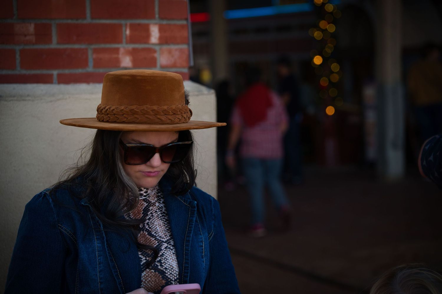 A person wearing a brown hat and sunglasses looks down while holding a drink.