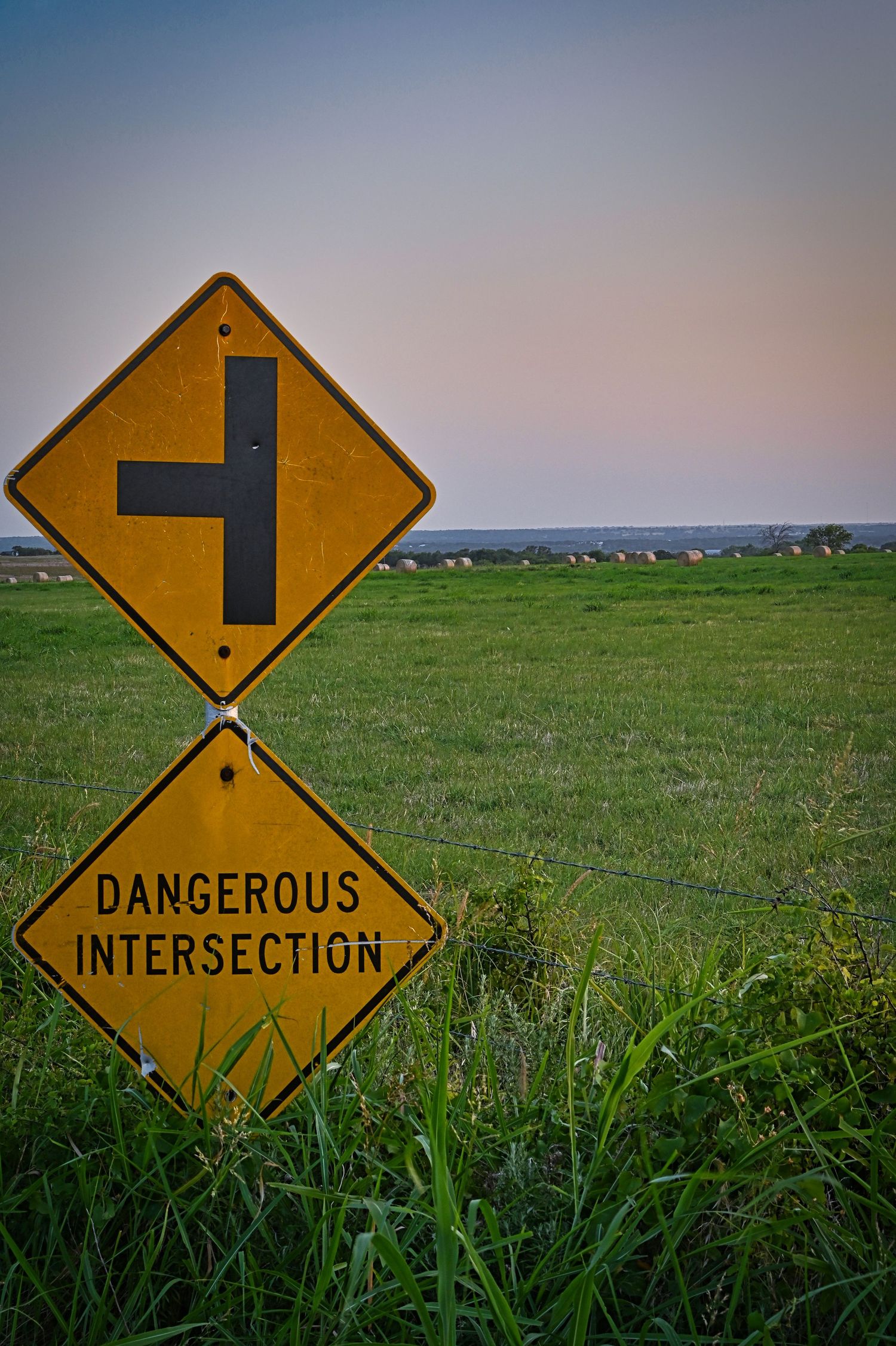 Yellow dangerous intersection warning sign in a rural field at dusk.