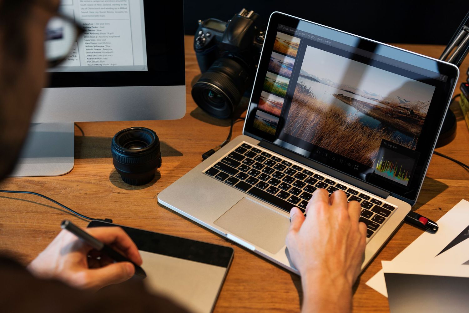 Person editing photos on laptop with camera and lens on wooden desk.