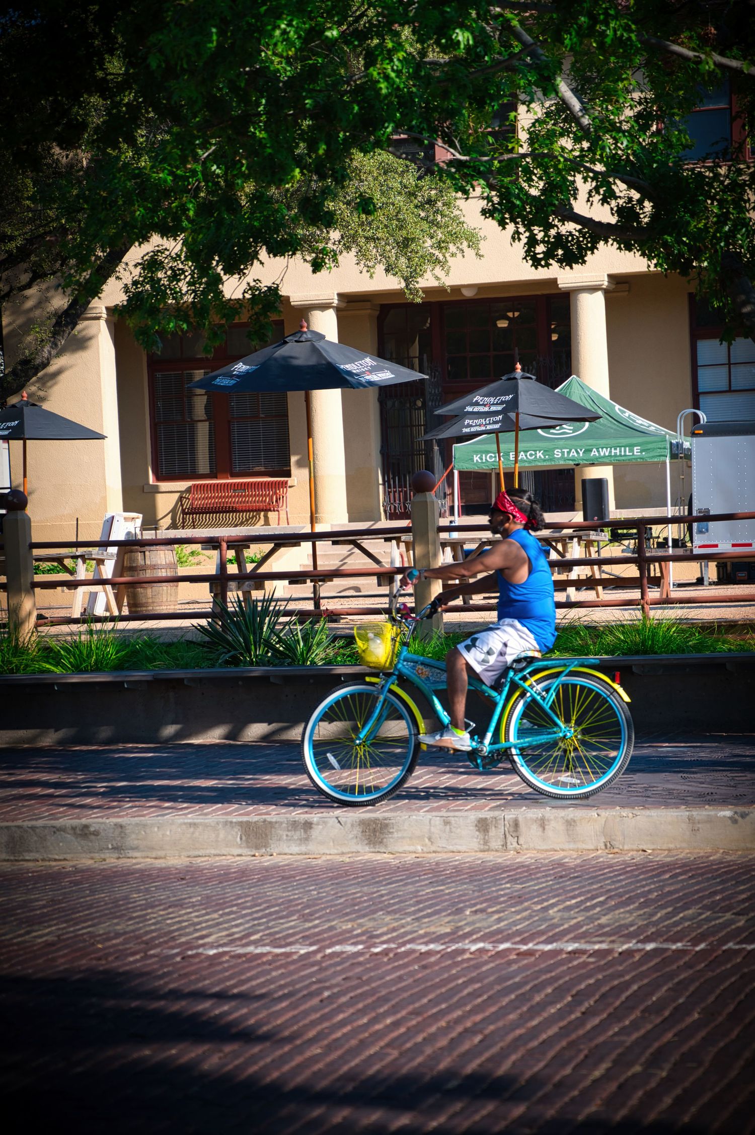 Cyclist riding bright turquoise bicycle past outdoor cafe with umbrellas and picnic tables.
