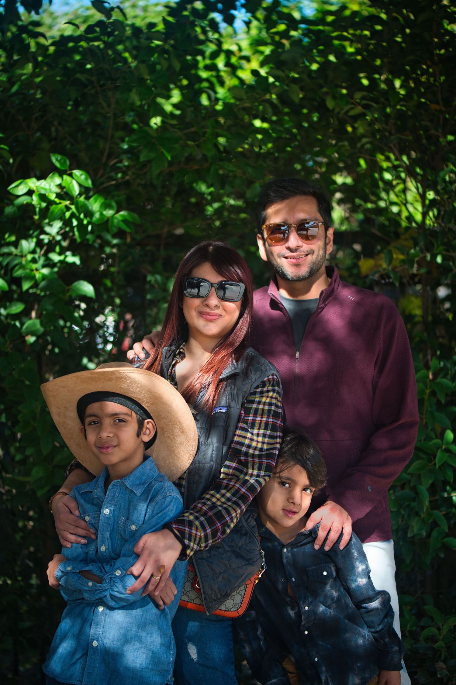 A family poses together outdoors in front of green foliage, wearing casual clothes and sunglasses on a sunny day.