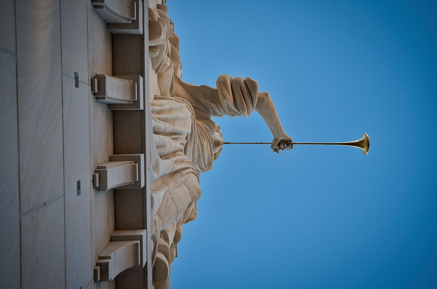 Architectural detail of a classical stone statue holding a trumpet against a bright blue sky.