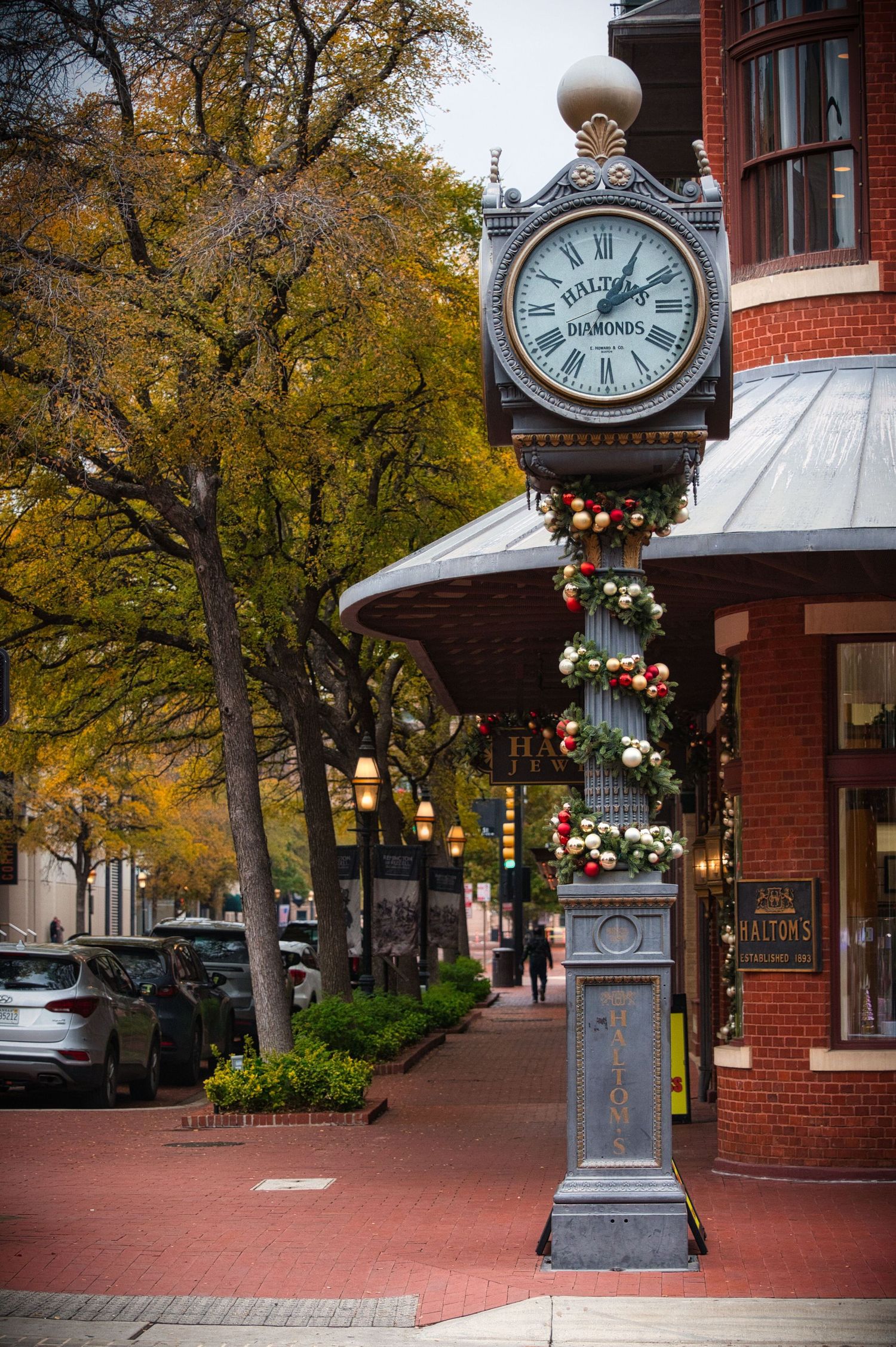 Decorative street clock adorned with holiday garland stands against autumn trees on brick sidewalk.