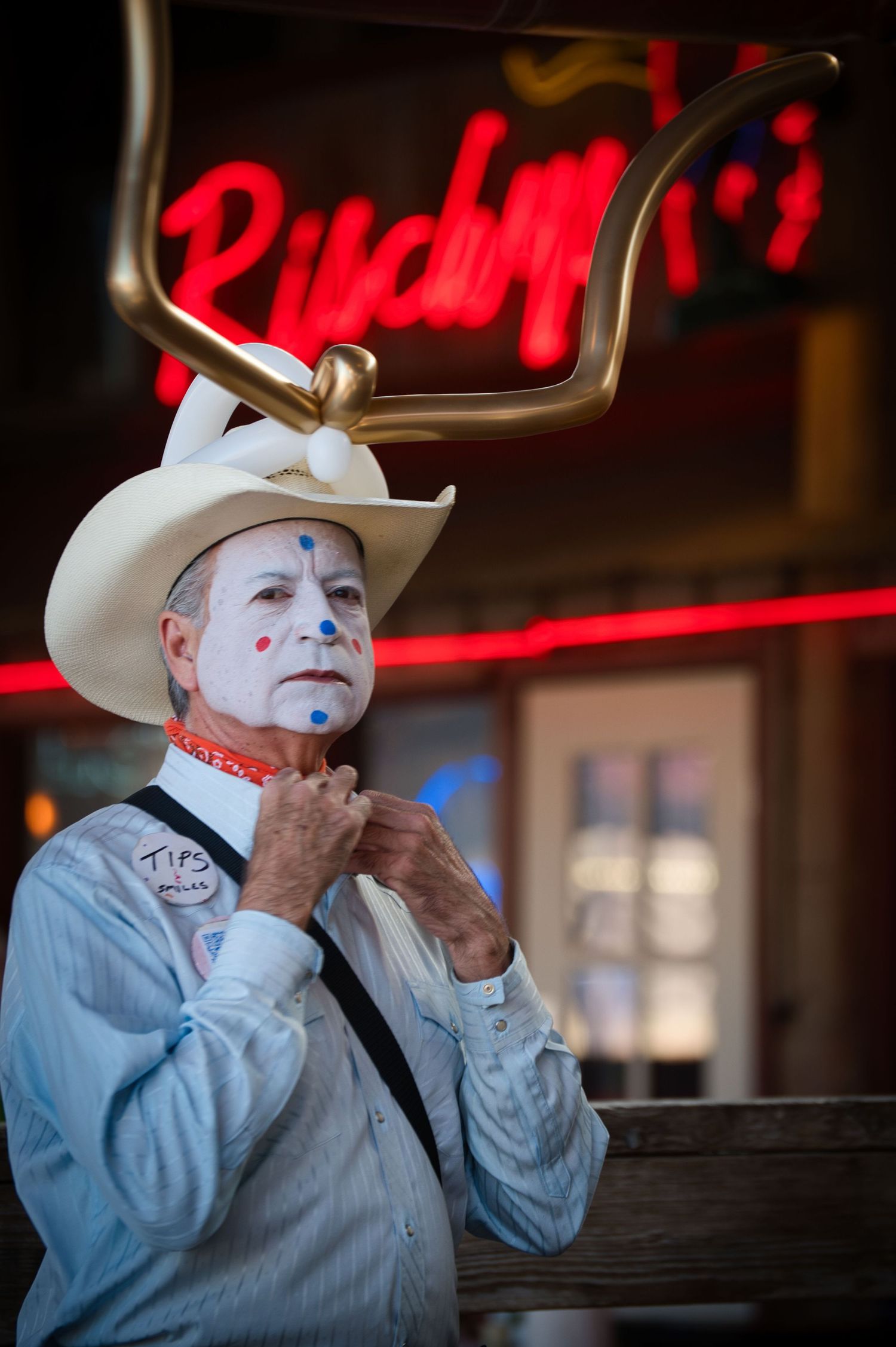 Performer in white cowboy hat and face paint poses under red neon lighting.