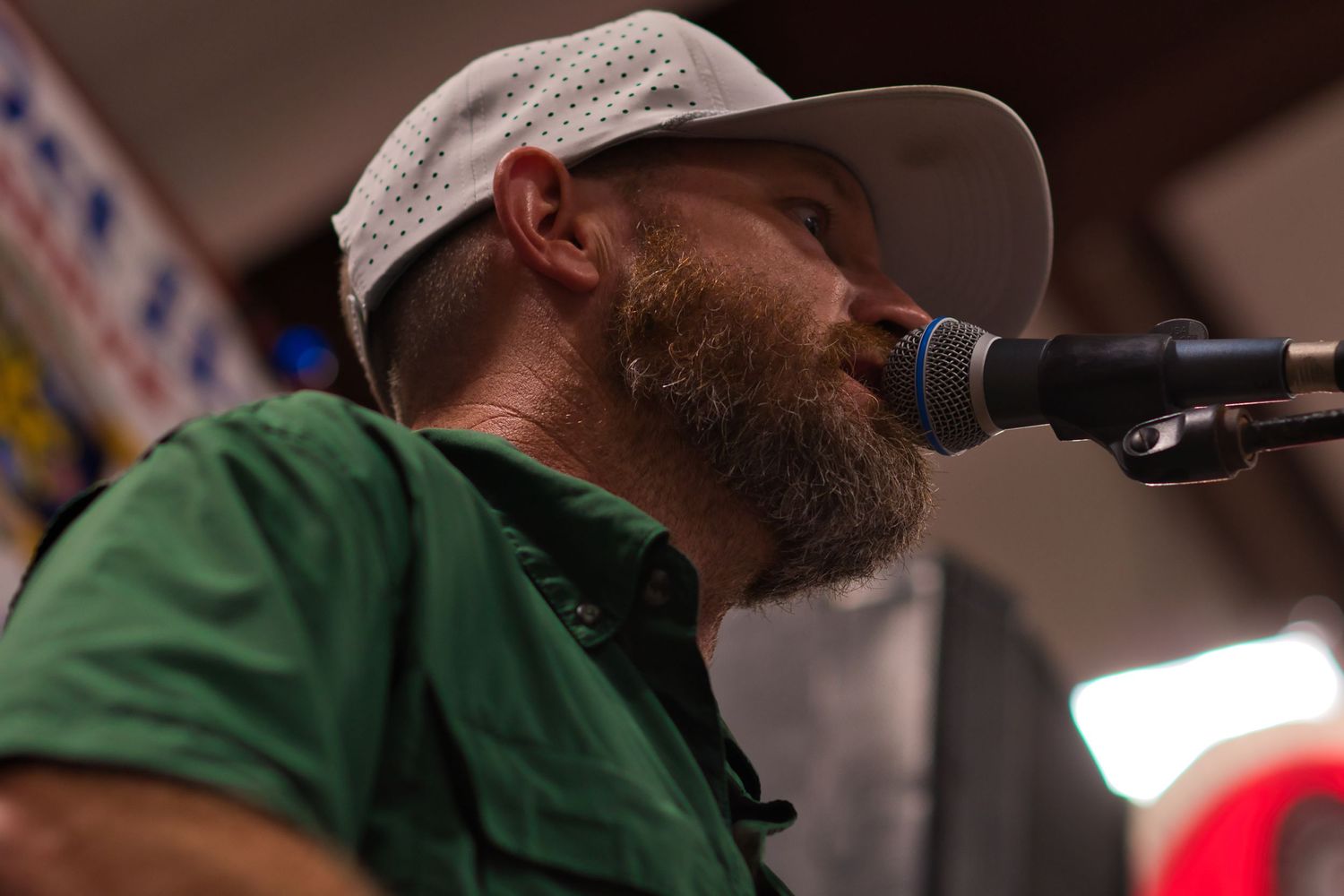 A bearded performer in a baseball cap speaks into a microphone on stage.