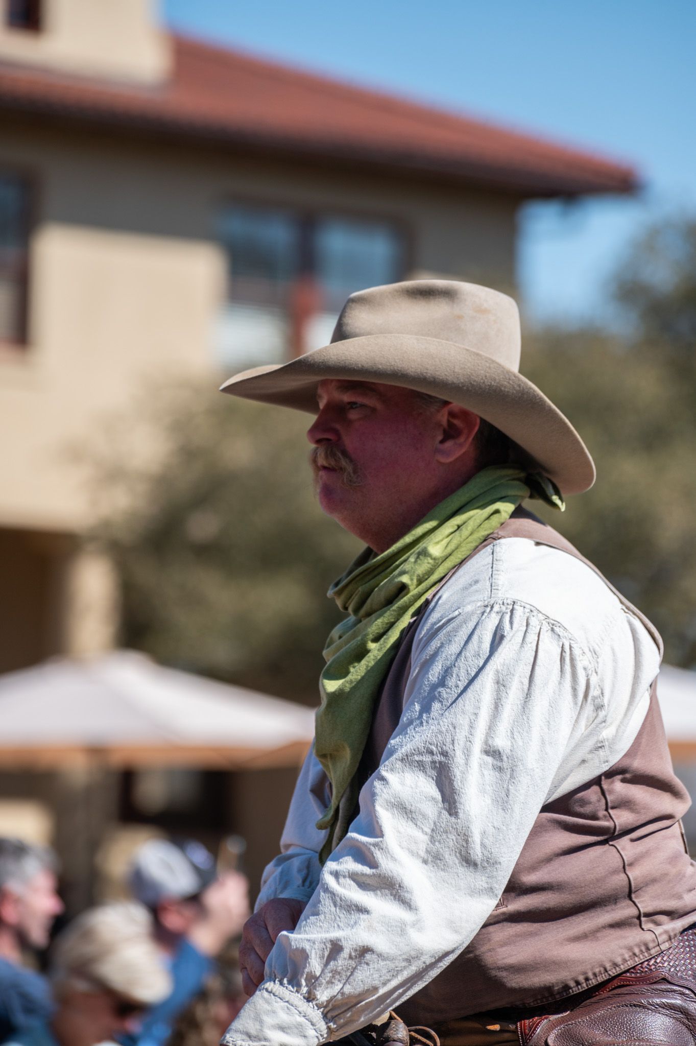 A person wearing a cowboy hat and vest with a green bandana stands at an outdoor western event.