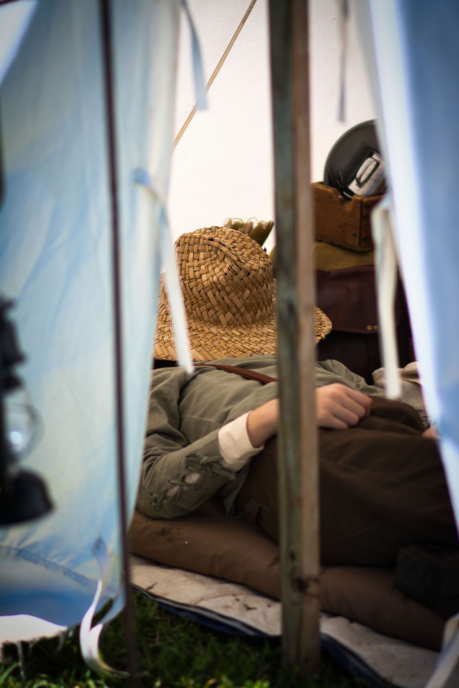 View through tent flaps of a War reenactor resting on bedding with a straw hat.