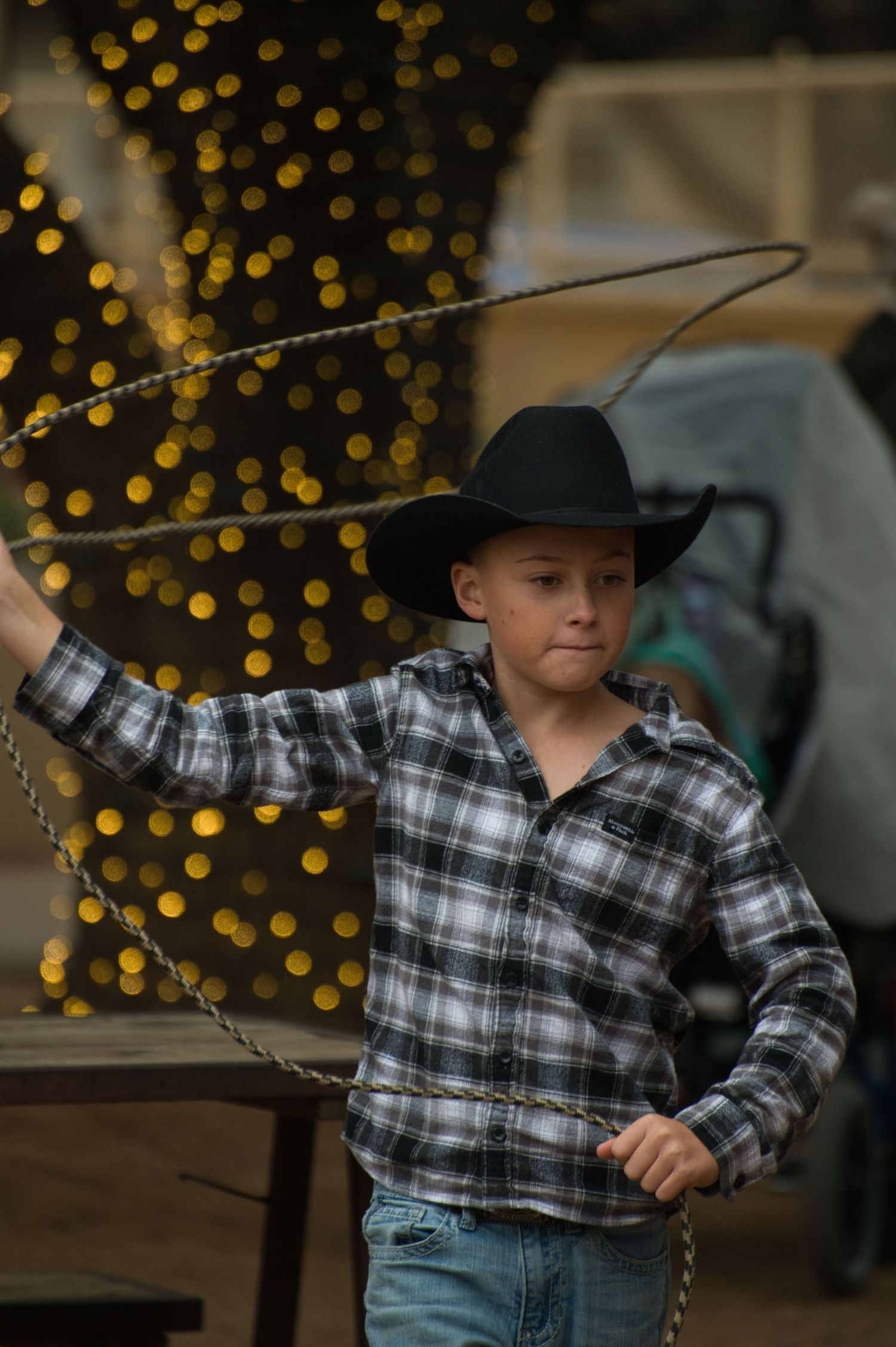 Young cowboy in plaid shirt and black hat stands against background of sparkly holiday lights.