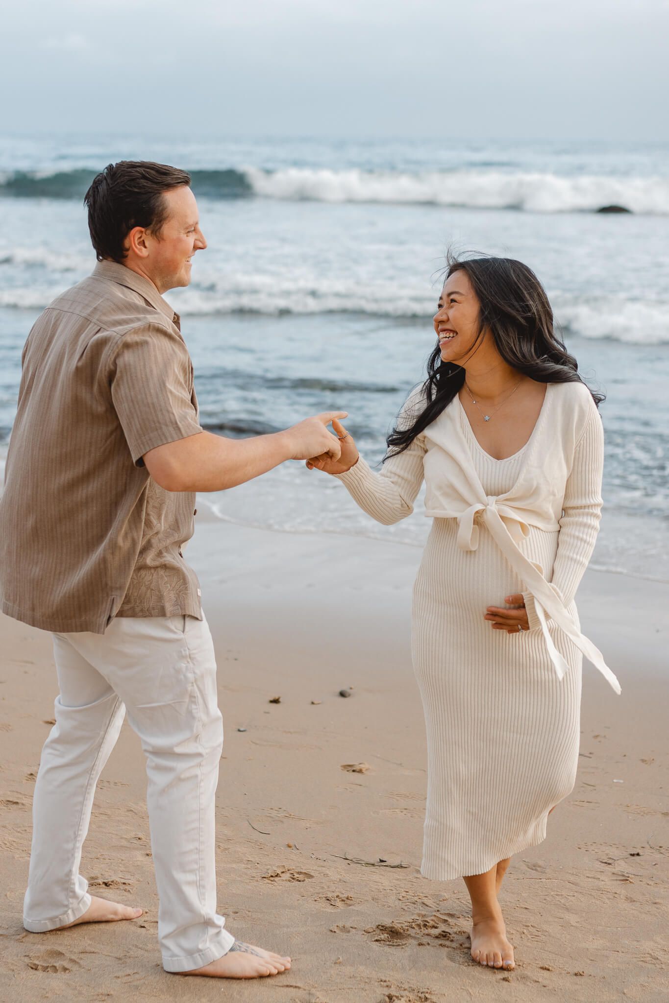 A happy couple holds hands on the beach while expecting a baby, with ocean waves in the background. Newport Pregnancy Shoot