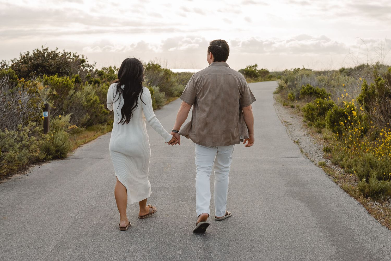 A couple walks hand in hand down a paved path surrounded by coastal vegetation at sunset. Newport Pregnancy Shoot