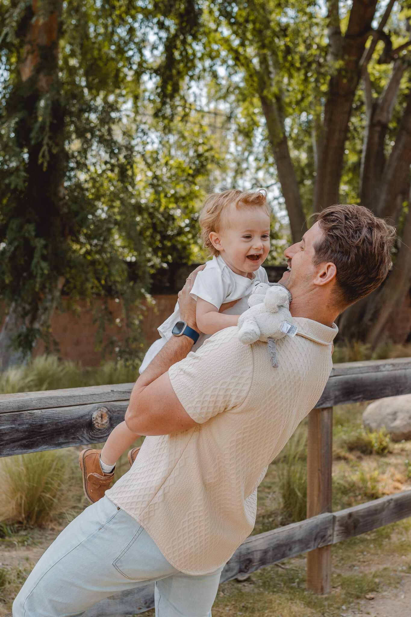 A tender outdoor moment captured in sunlit woods as parent and toddler share joyful interaction by a wooden fence.
