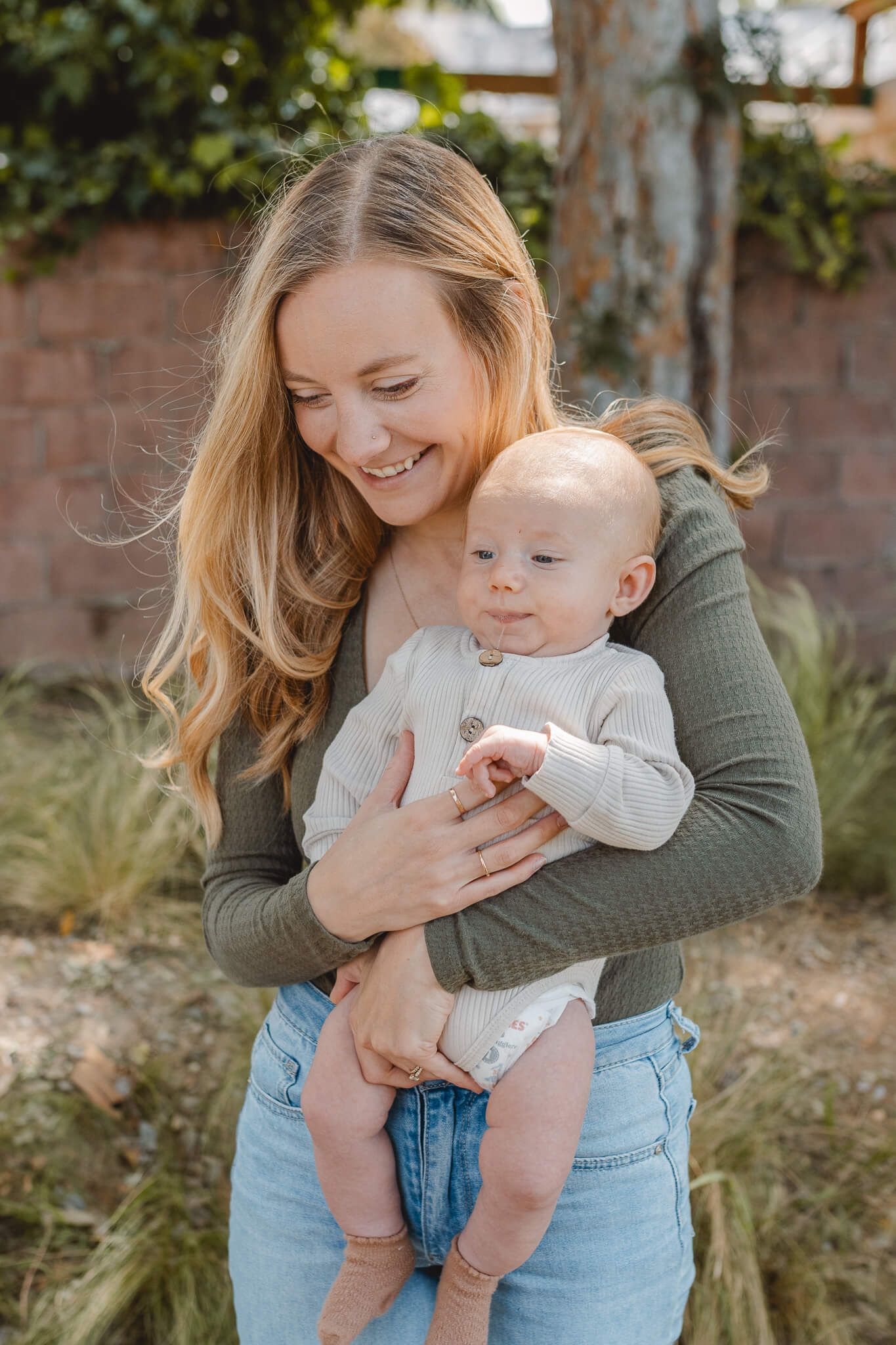 A joyful mother in olive green sweater holds and smiles at her baby wearing a white onesie in an outdoor setting.