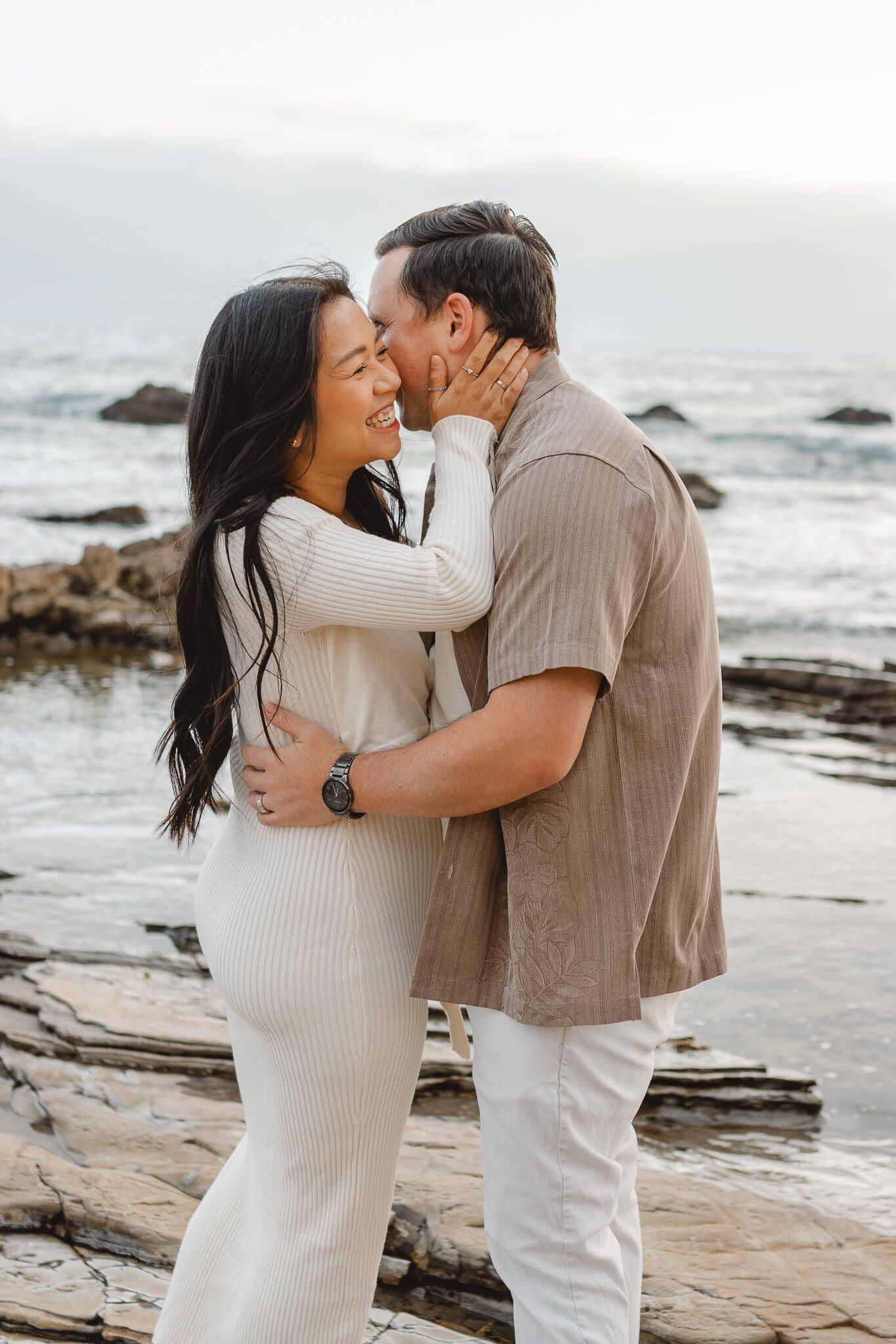 A romantic couple embraces on a rocky beach at sunset wearing coordinating neutral-colored outfits. Newport Pregnancy Shoot