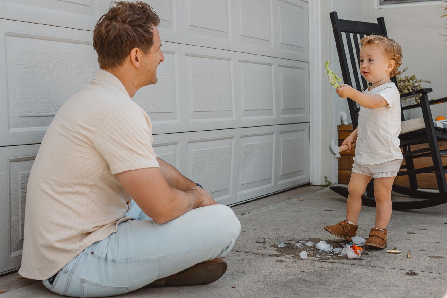 A toddler in white clothing stands near a rocking chair while someone sits on the floor of a garage.