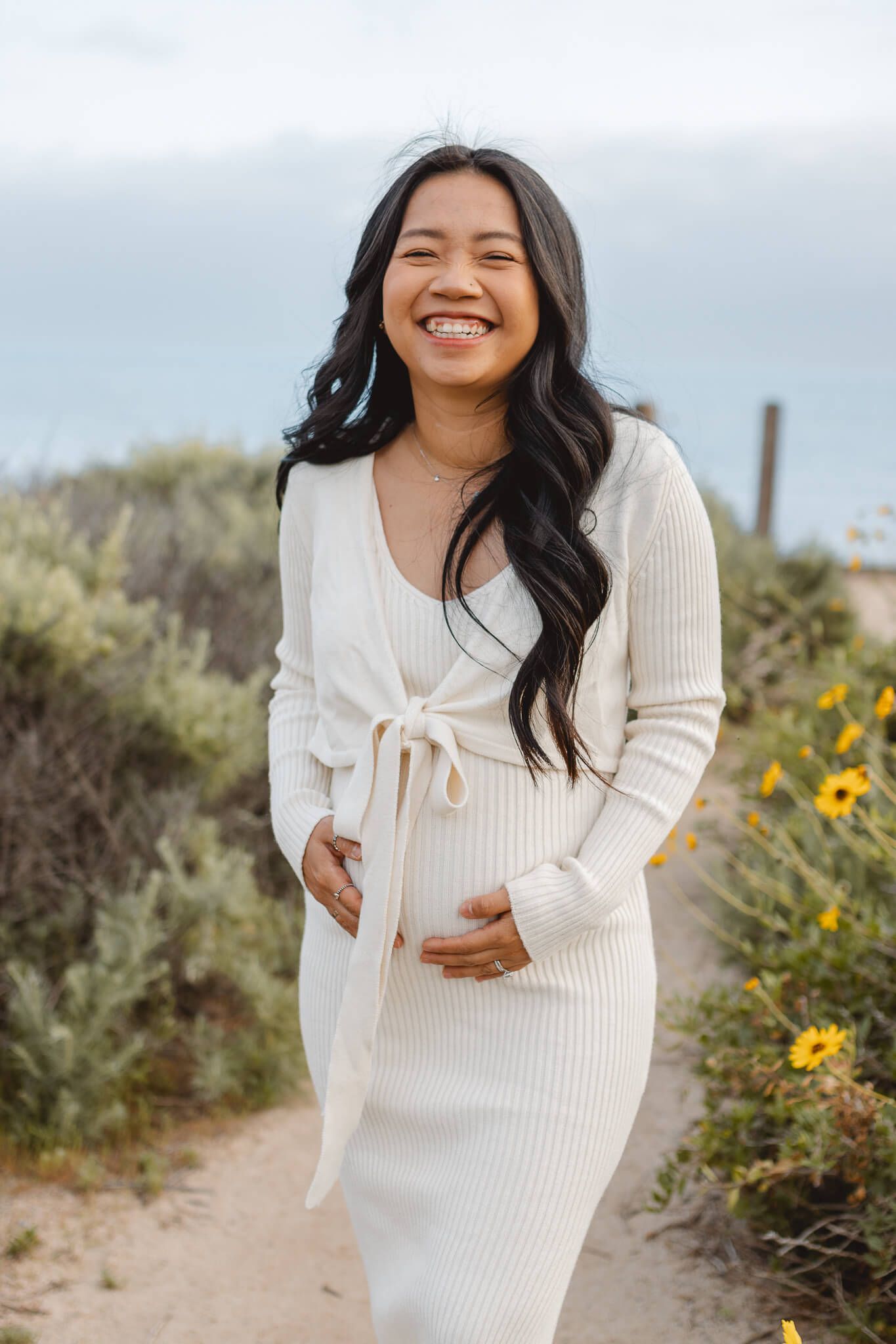 A person wearing a cream ribbed knit wrap dress stands smiling on a nature path with yellow wildflowers in the background. Newport Pregnancy