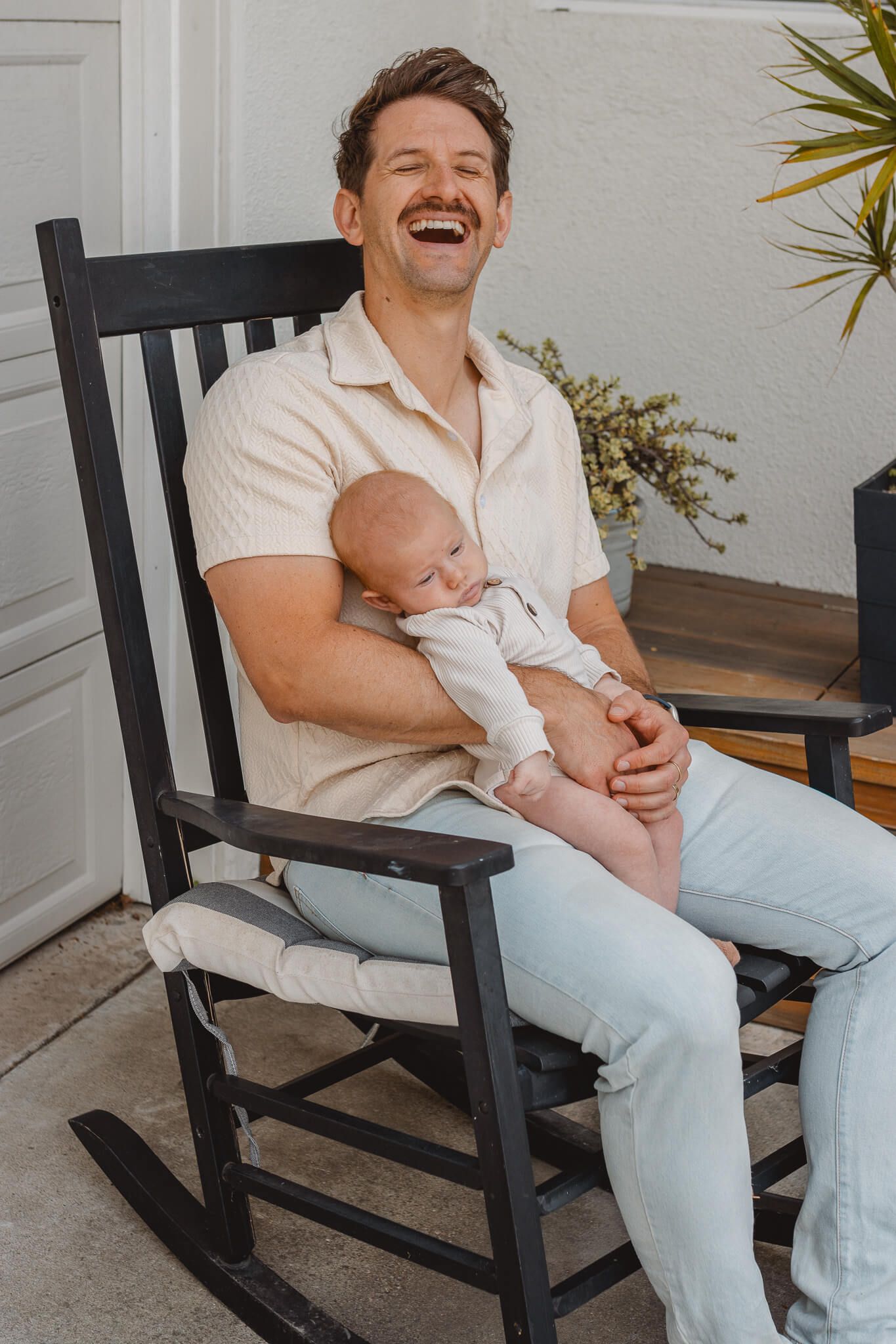 A parent and baby share a joyful moment while sitting in a black rocking chair on a porch.