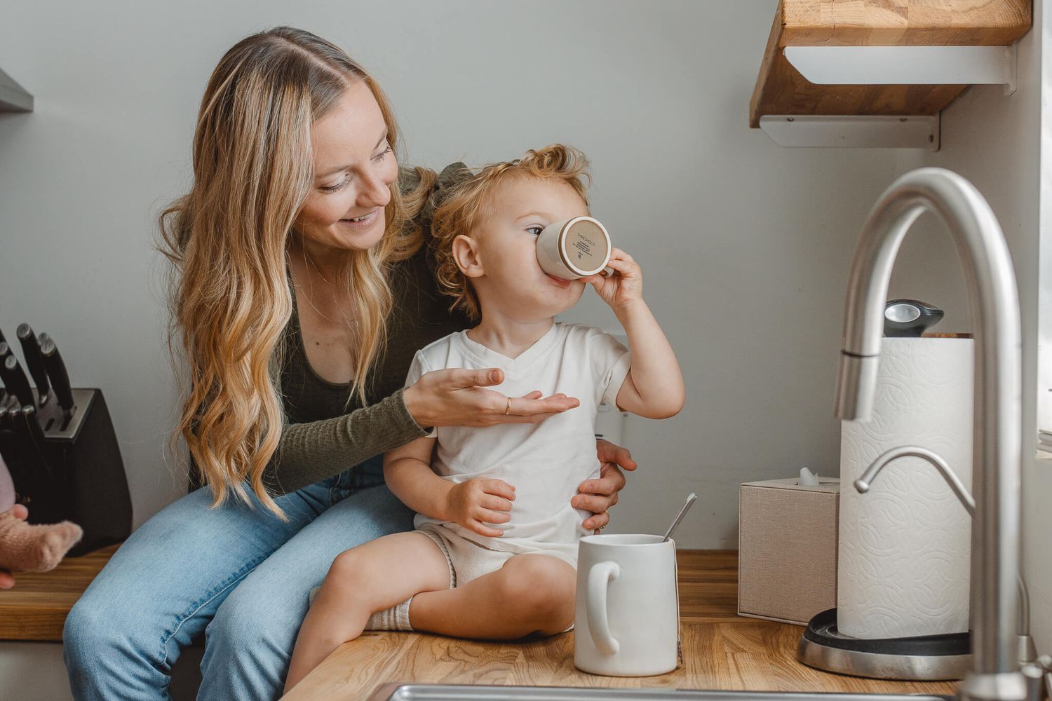 A warm kitchen scene shows a child sipping from a cup while sitting on the counter with someone in casual attire.