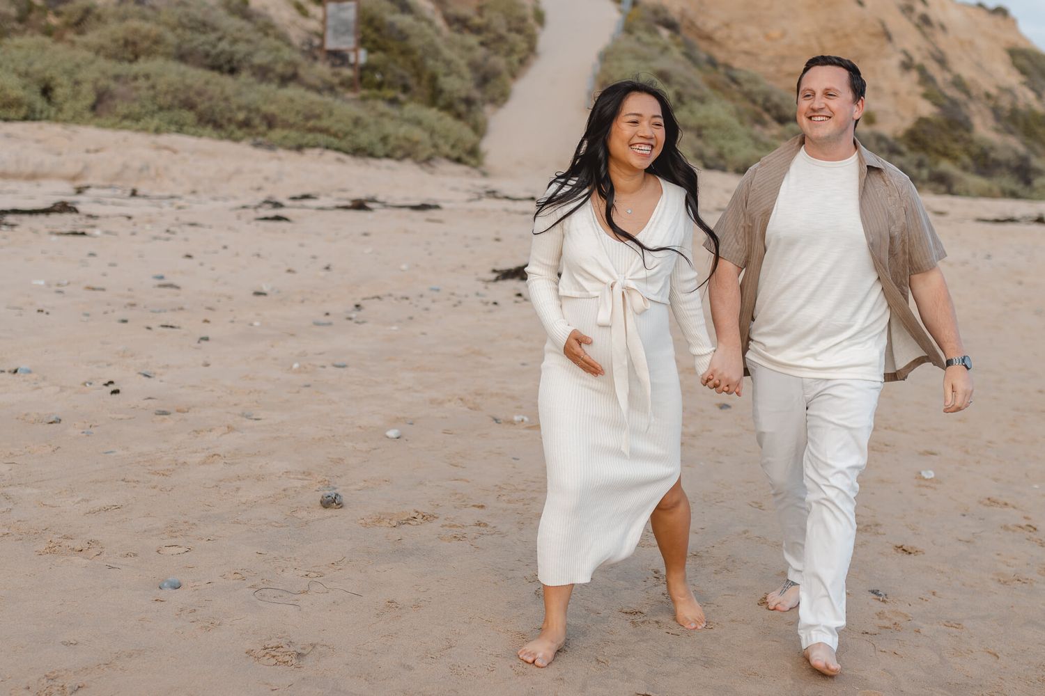 A couple walks hand in hand on a sandy beach near cliffs during sunset, both dressed in white casual clothing. Newport Pregnancy Shoot
