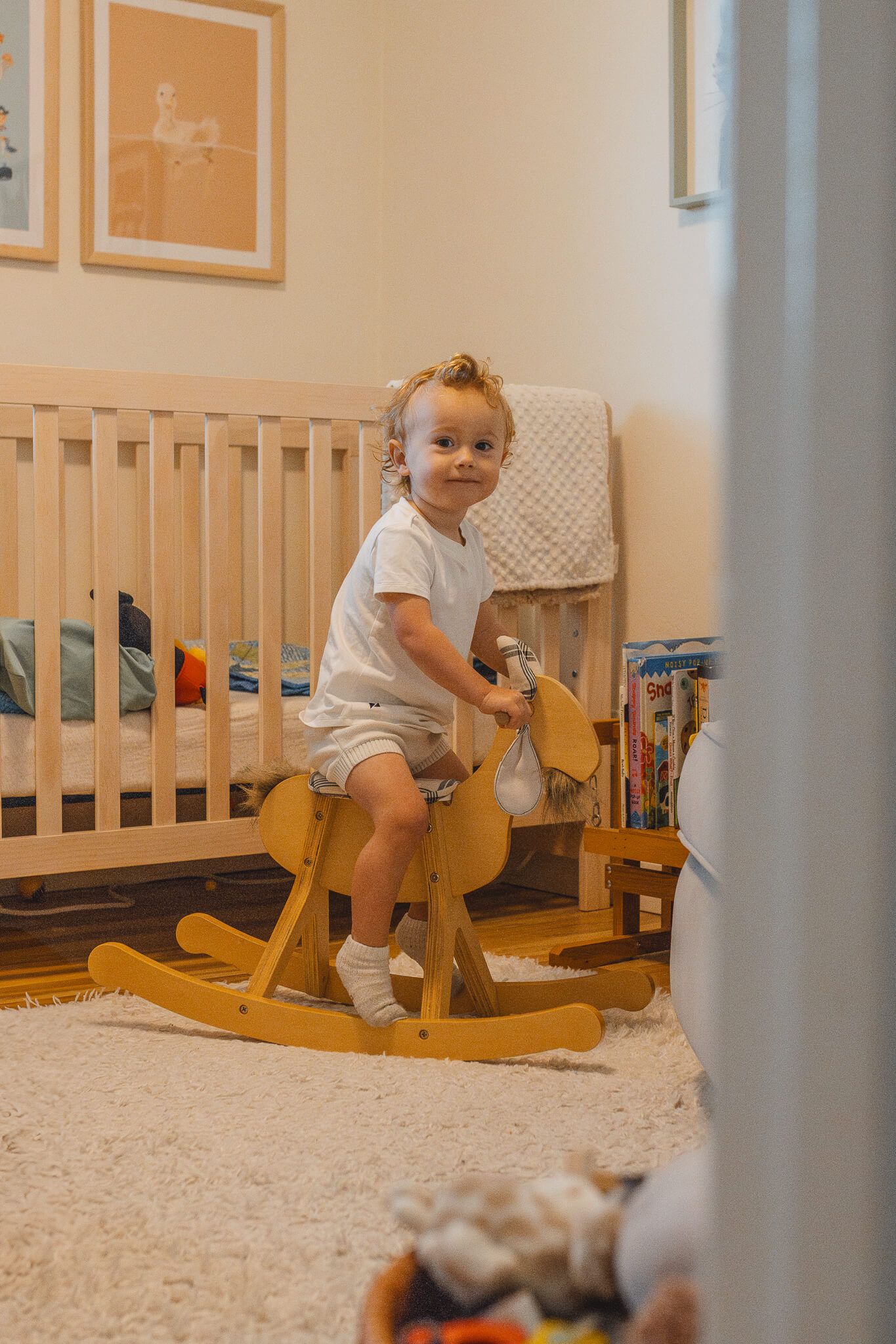 A toddler rides a wooden rocking horse in a room with cream carpet and framed wall art.