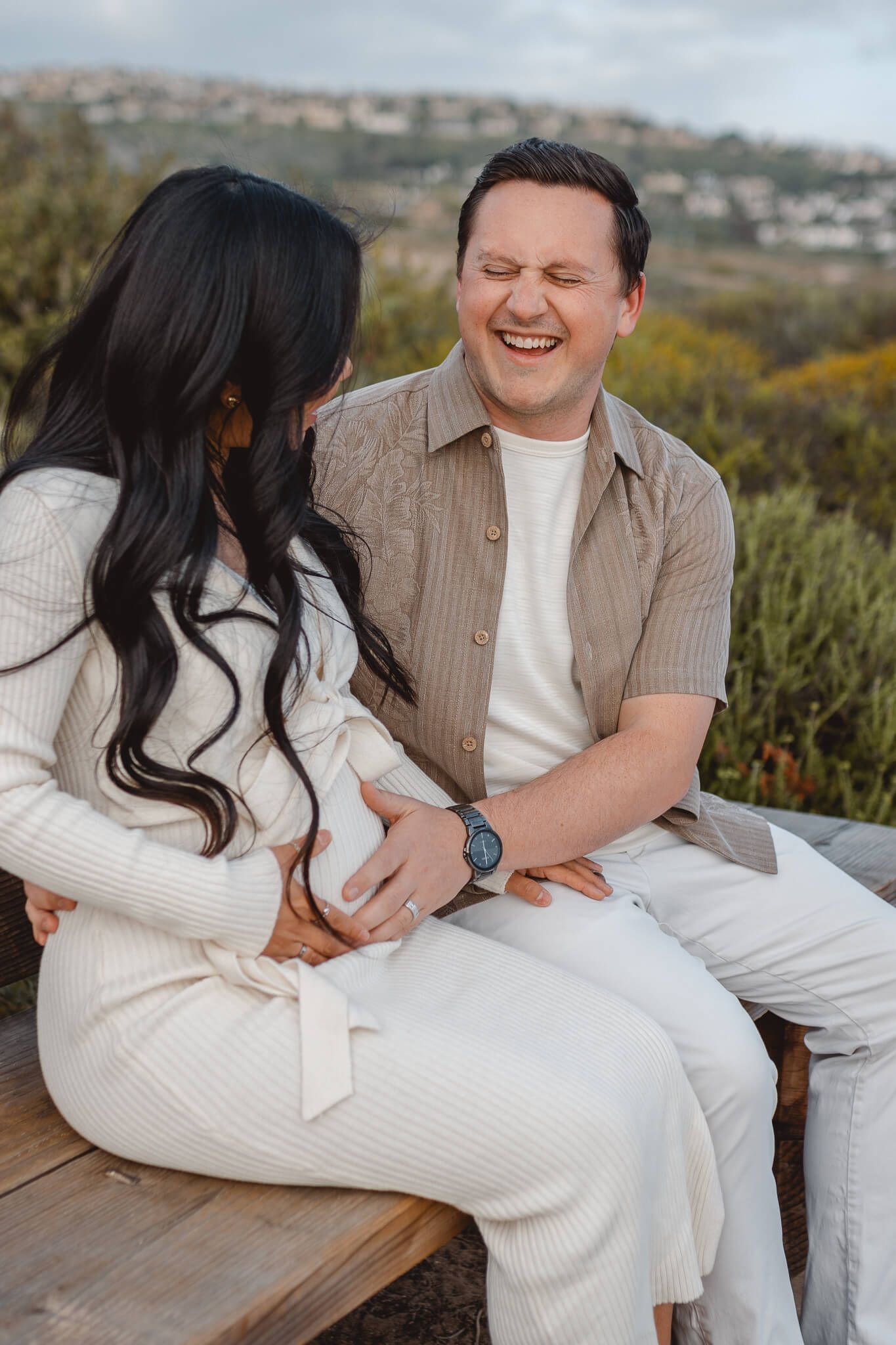 Couple in white and beige outfits sharing a joyful moment while sitting on a wooden bench against a hillside landscape. Newport Pregnancy