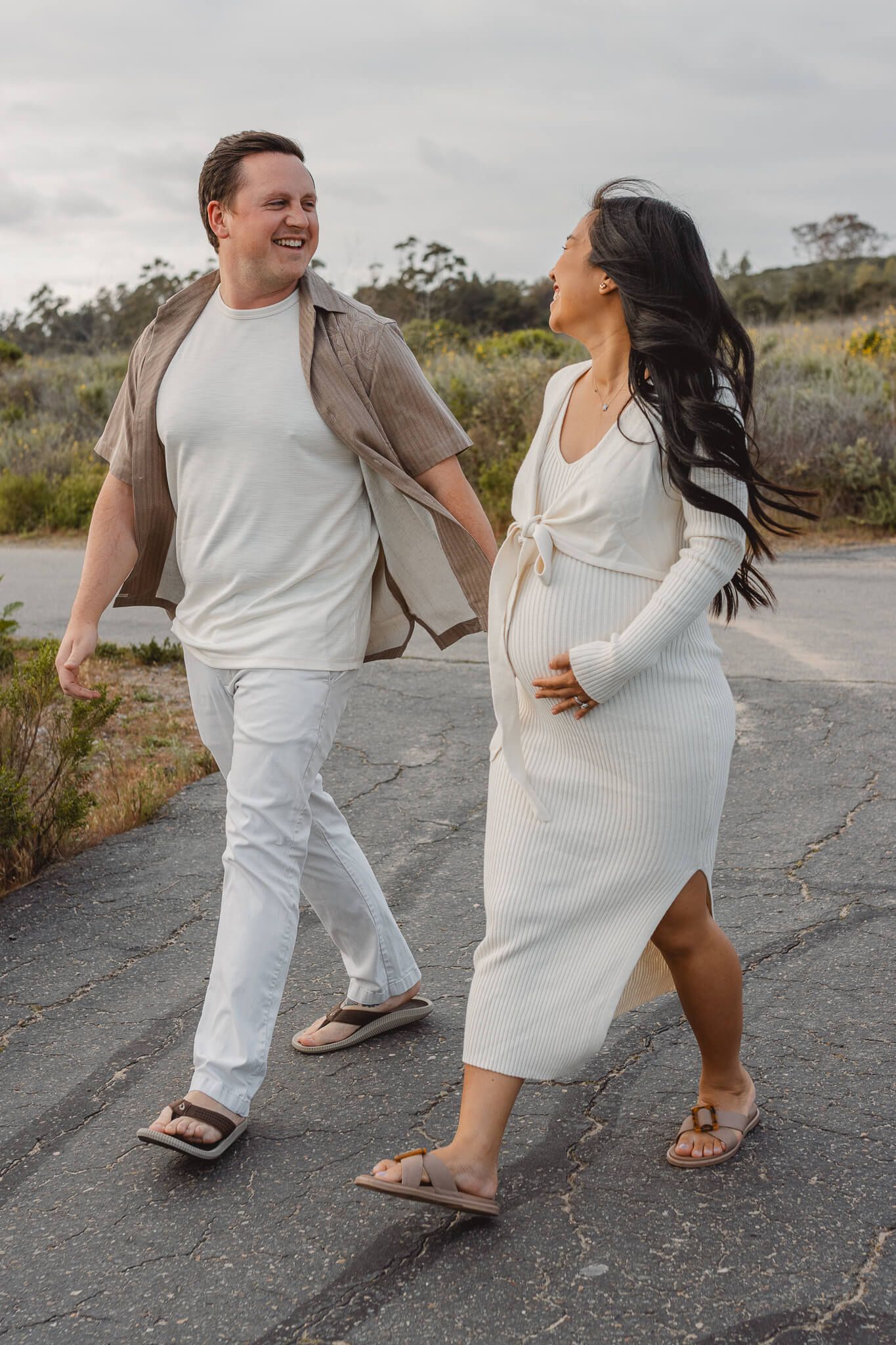 A couple walking together on a path wearing light colored outfits, with the woman in a fitted white dress and sandals. Newport Pregnancy