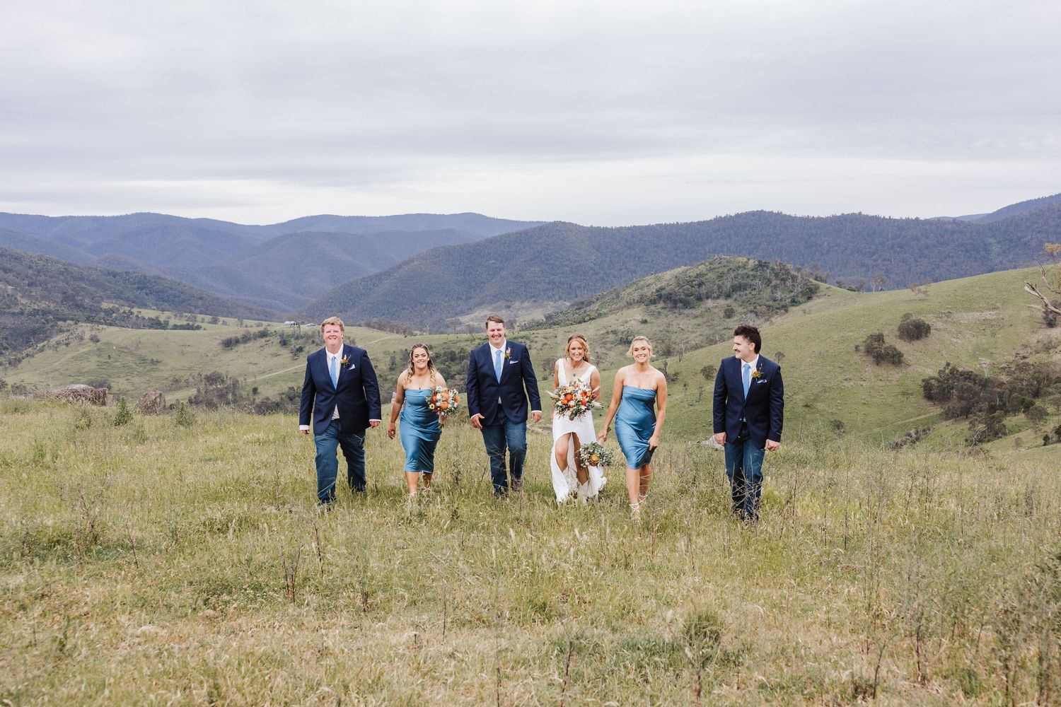 Wedding party walks together across a grassy hillside with scenic mountain ranges in the background on an overcast day.