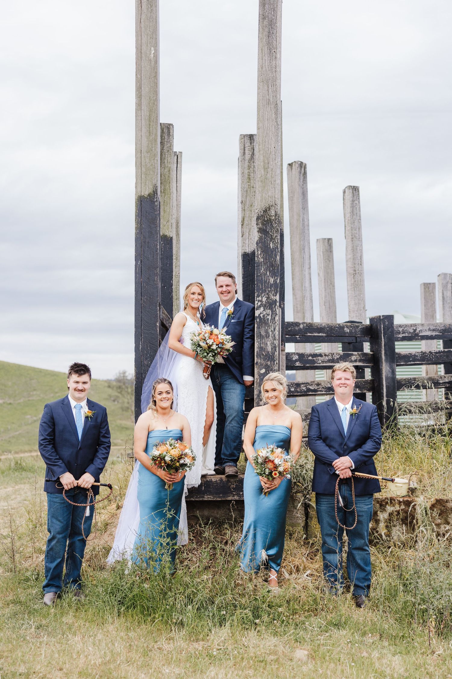 Wedding party poses in front of rustic wooden posts in a field with light blue bridesmaid dresses and navy suits.