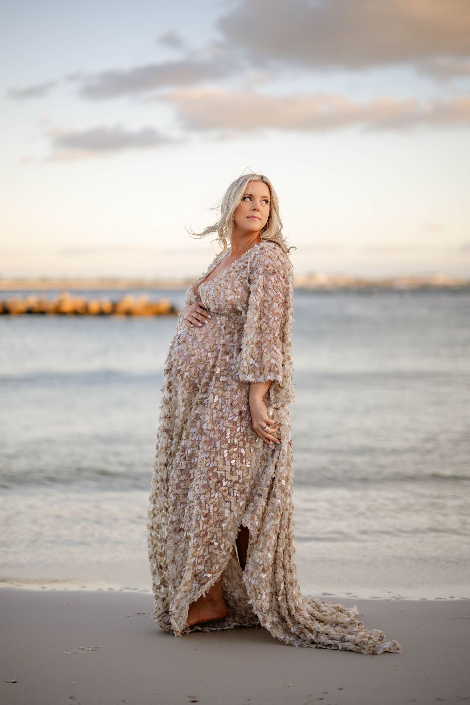 Expectant mother in flowing beige dress poses on beach at golden hour.