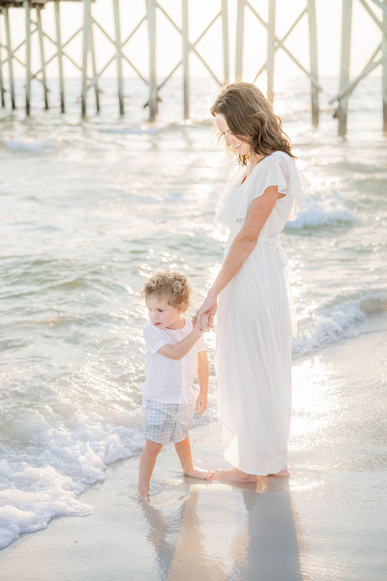 A white-dressed person holding hands with a toddler while walking along the beach shore near a pier at sunset.