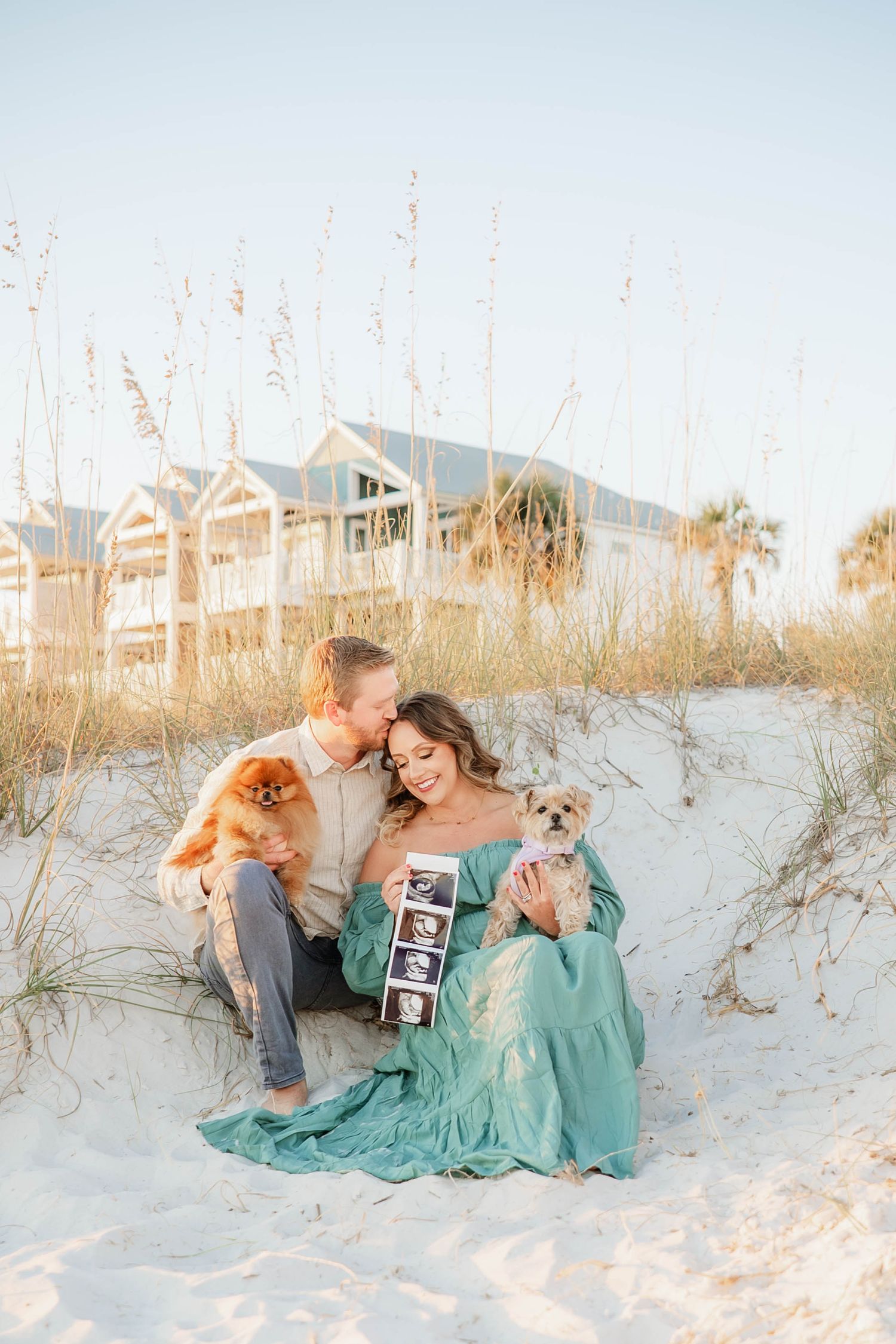 A couple sits on a beach sand dune with two dogs while holding ultrasound photos at sunset near coastal homes.