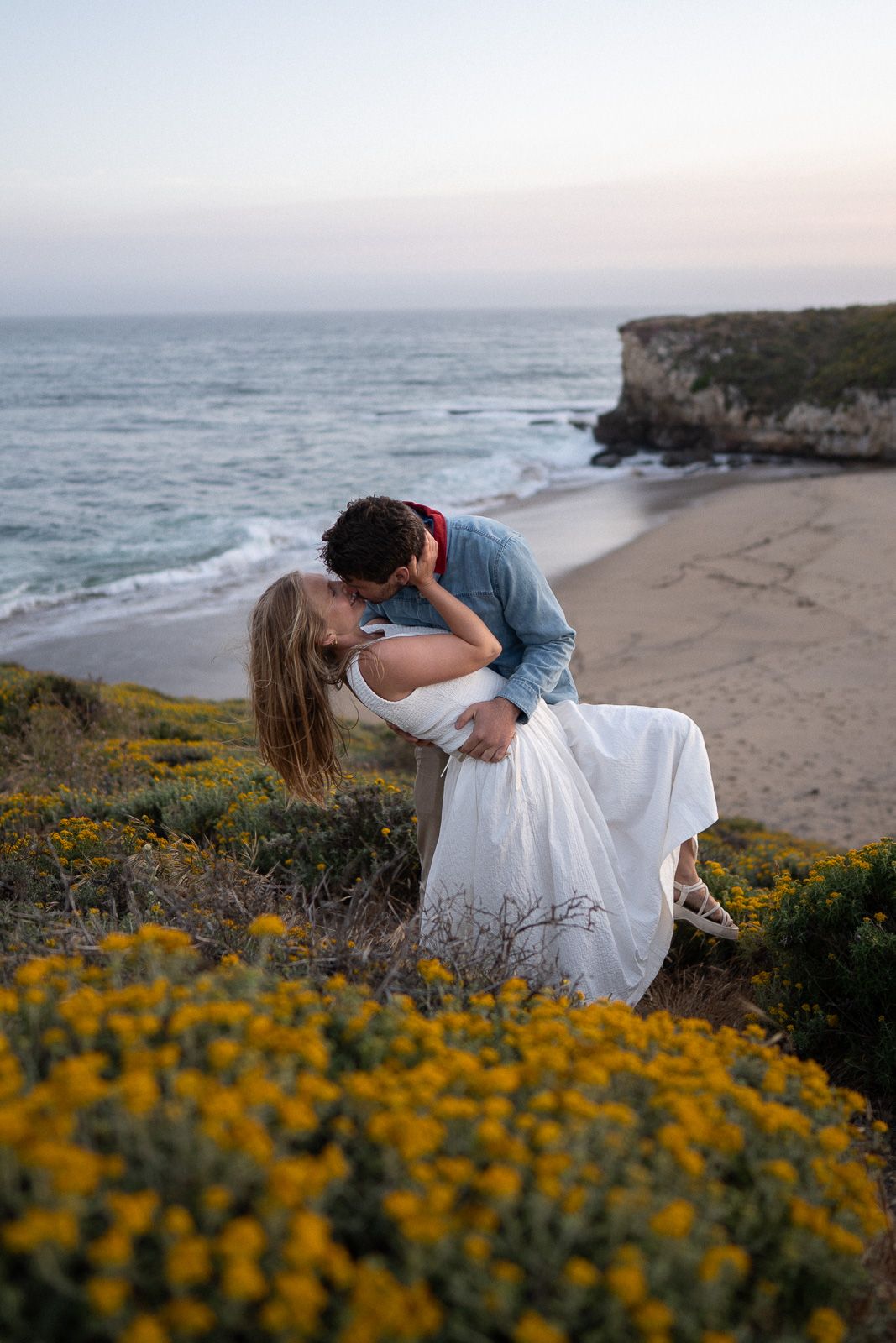 Dreamy coastal scene with figures in white dresses dancing among yellow wildflowers on a clifftop overlooking the ocean.