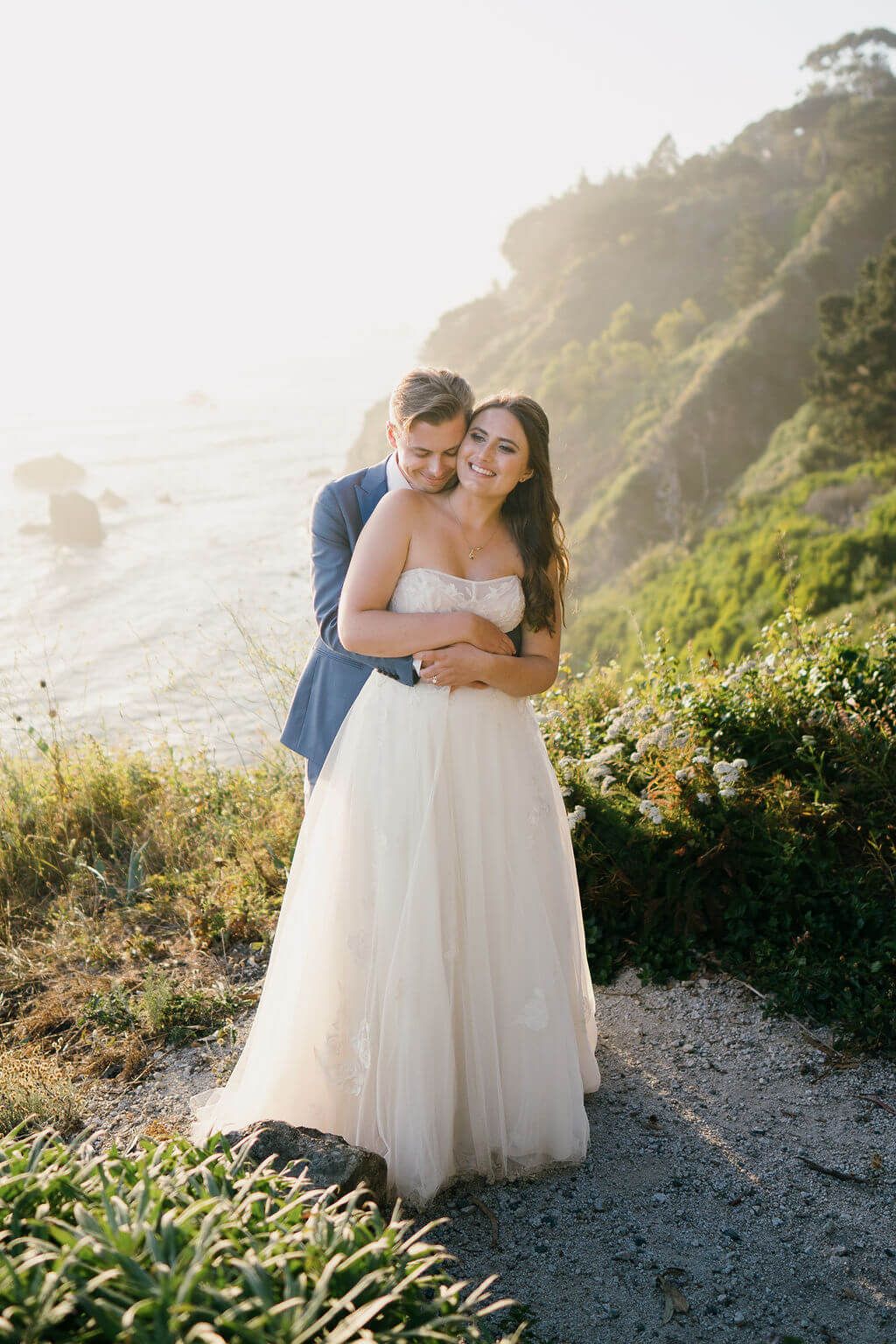 A romantic couple in formal attire embraces along a scenic coastal cliff during golden hour at sunset.