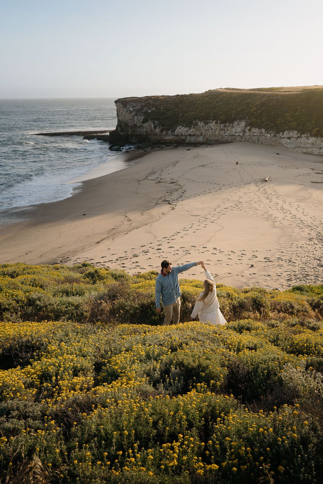 Two silhouetted figures stand on a coastal cliff overlooking a sandy beach with white cliffs and yellow wildflowers at sunset.