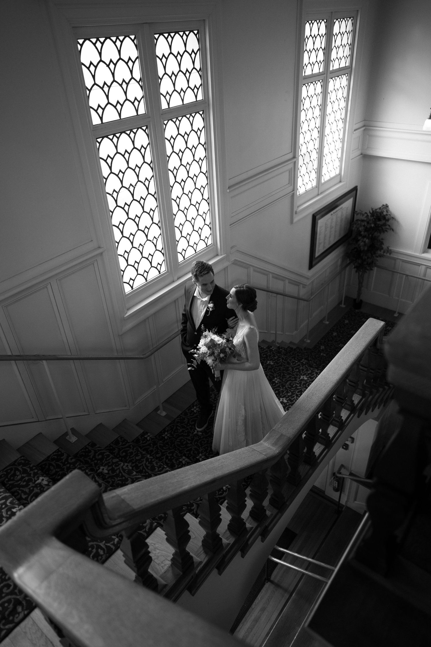 A black and white photo capturing a romantic stairway moment with gothic windows casting light in an elegant interior.