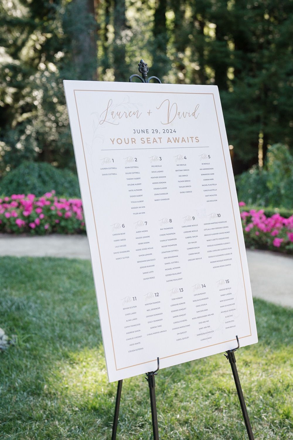 An elegant white wedding seating chart displayed on an easel outdoors with pink flowers and greenery in the background.