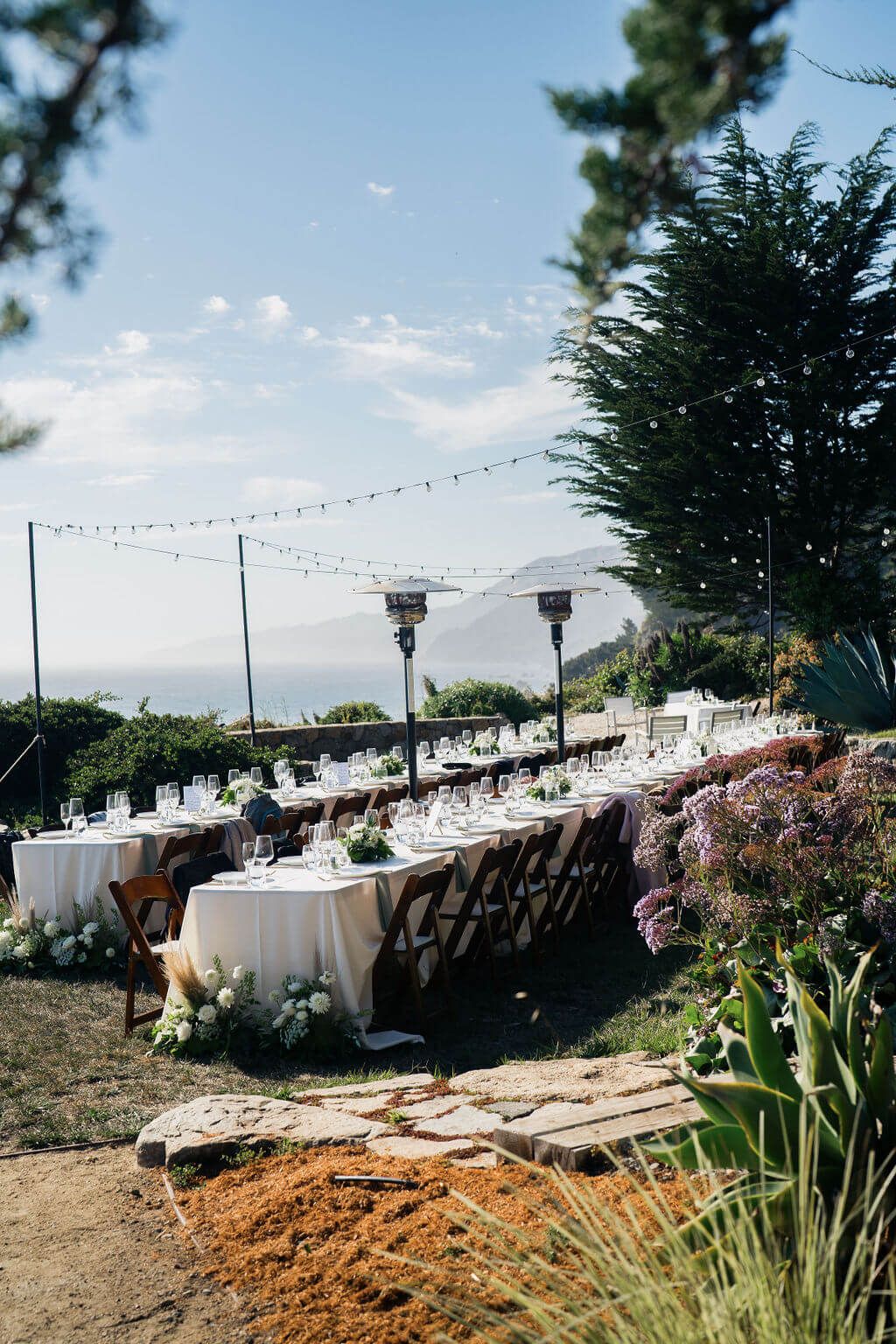 Outdoor beach wedding setup with long banquet tables, white linens, and string lights overlooking the ocean at sunset.
