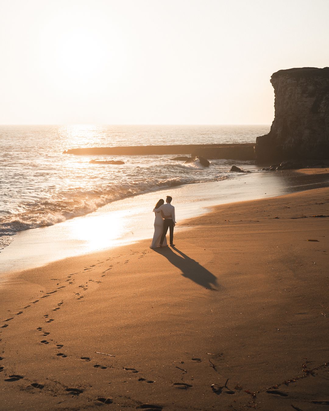 Silhouettes cast long shadows on a beach at sunset with footprints in the sand and dramatic coastal cliffs in the background.