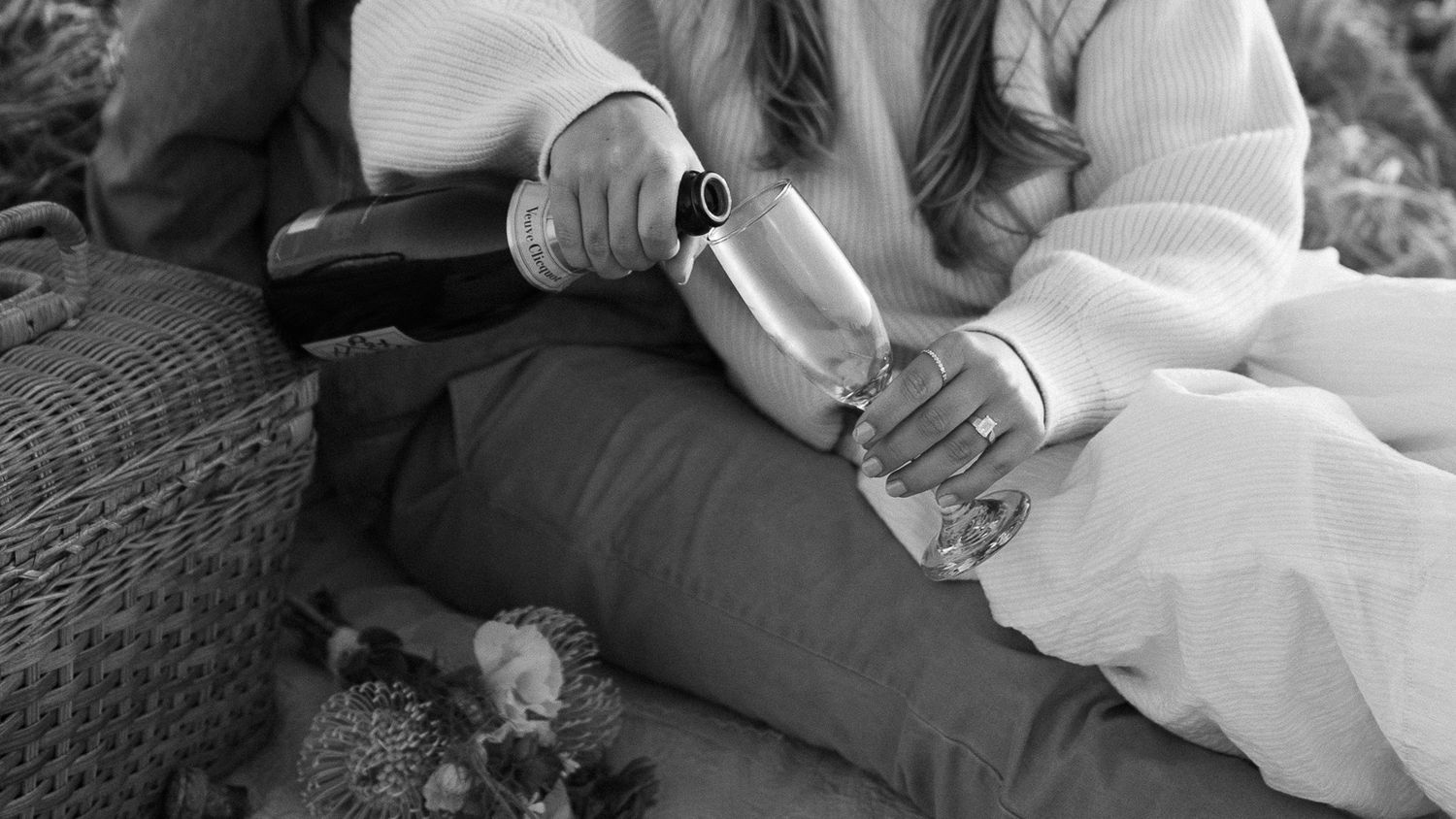 A black and white close-up shot of hands pouring a liquid drink into a cup while sitting outdoors.