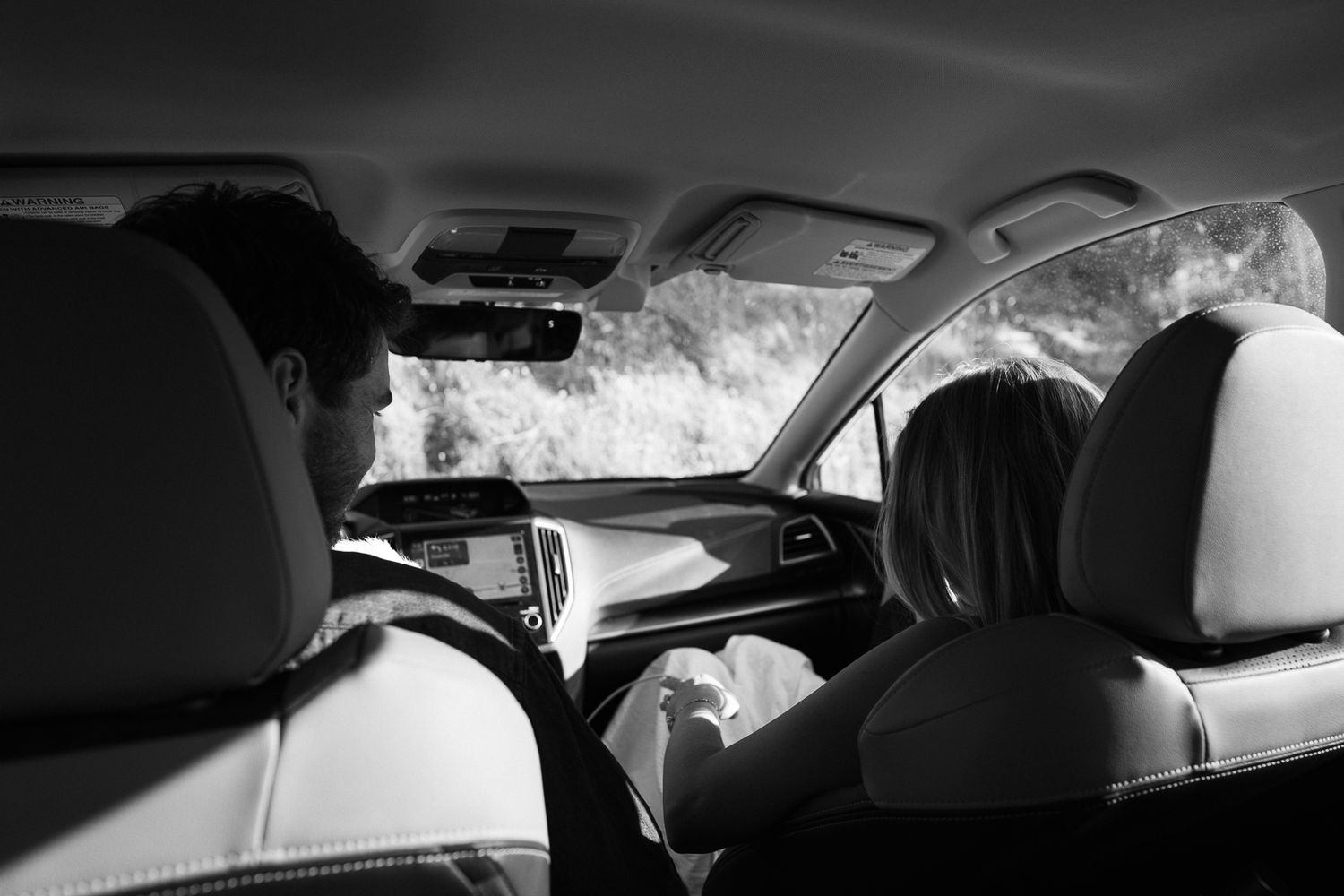 Black and white interior view of car from backseat showing front seats, headrests and windshield during daytime drive.